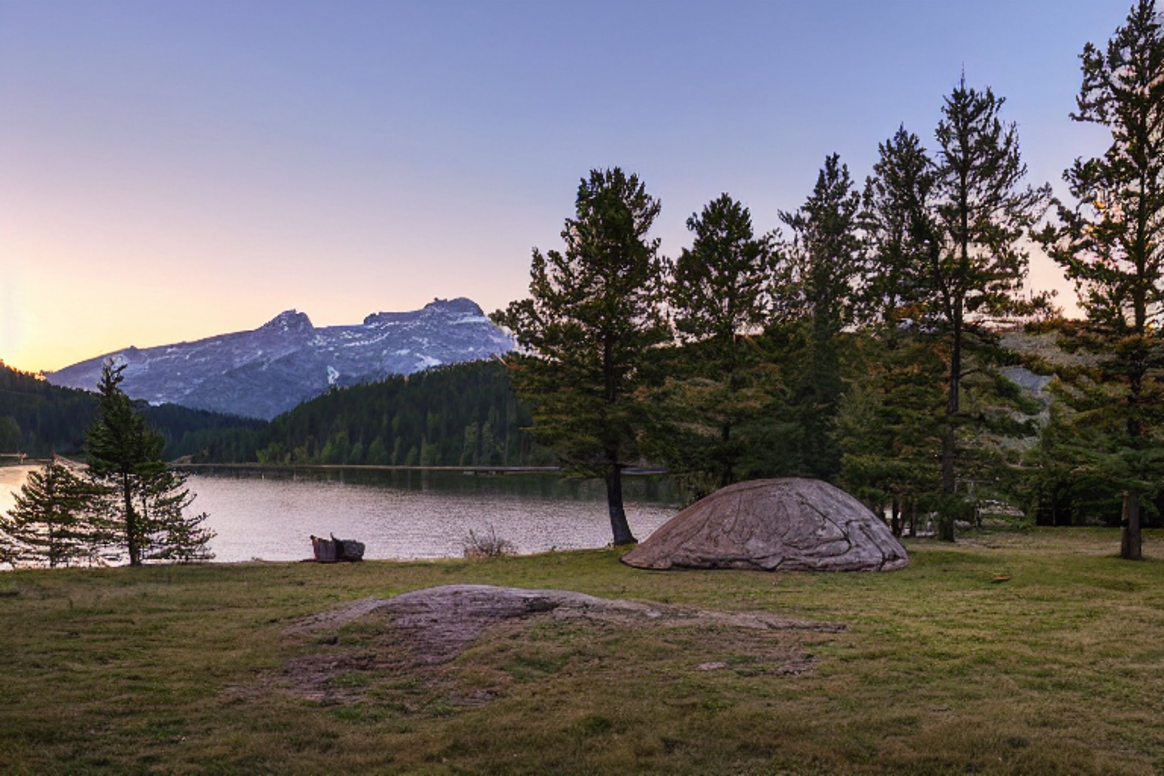 A brown tent rests on a paved campsite beside a peaceful lake. Golden sunlight warms the small trees, large rocks, and dirt clearing. Tall trees frame the view, with a mountain rising in the distance.