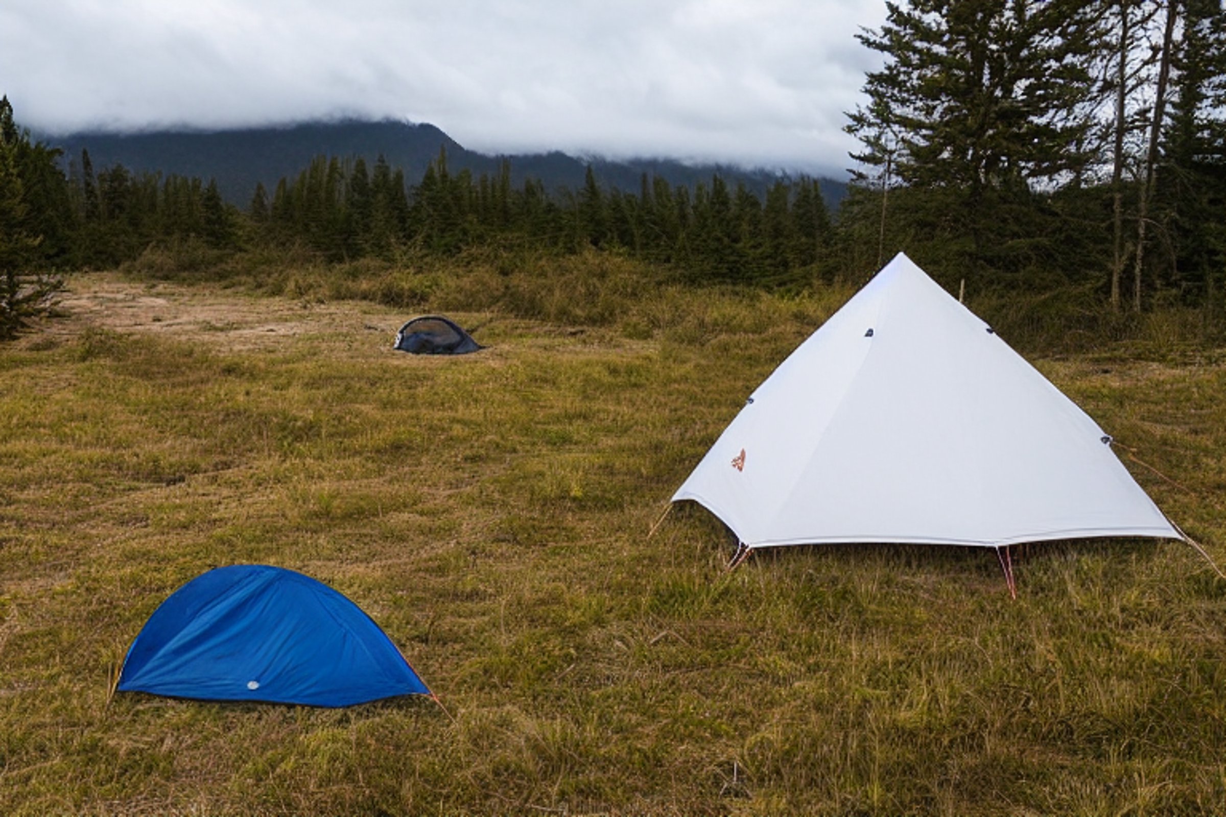 A white tent rests beside a glowing campfire on a quiet beach. Tall grass and rough ground surround the campsite, with camping chairs nearby. Dark mountains rise in the distance beneath the cloudy night sky.