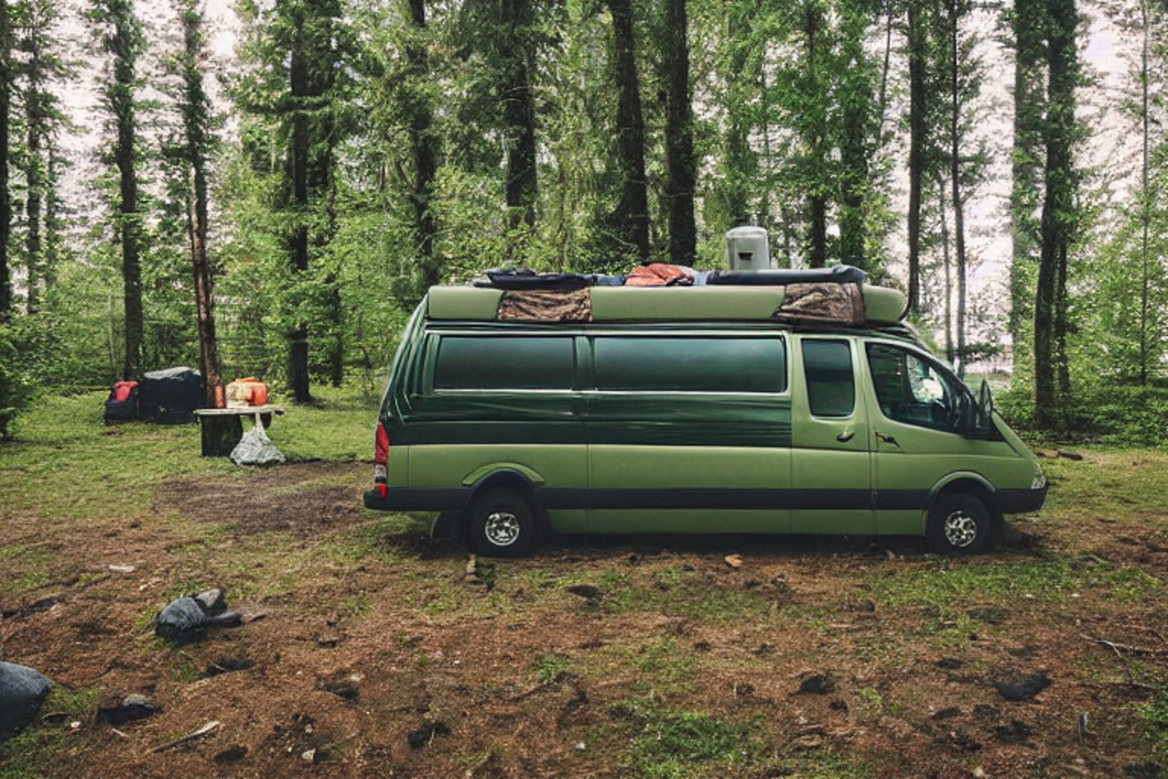 A green-and-tan camper van rests among small trees in a quiet Washington campsite. A warm campfire glows beside the van, while a lantern and backpack sit on the gravelly ground. Rainy clouds drift beneath a bright, star-filled sky.