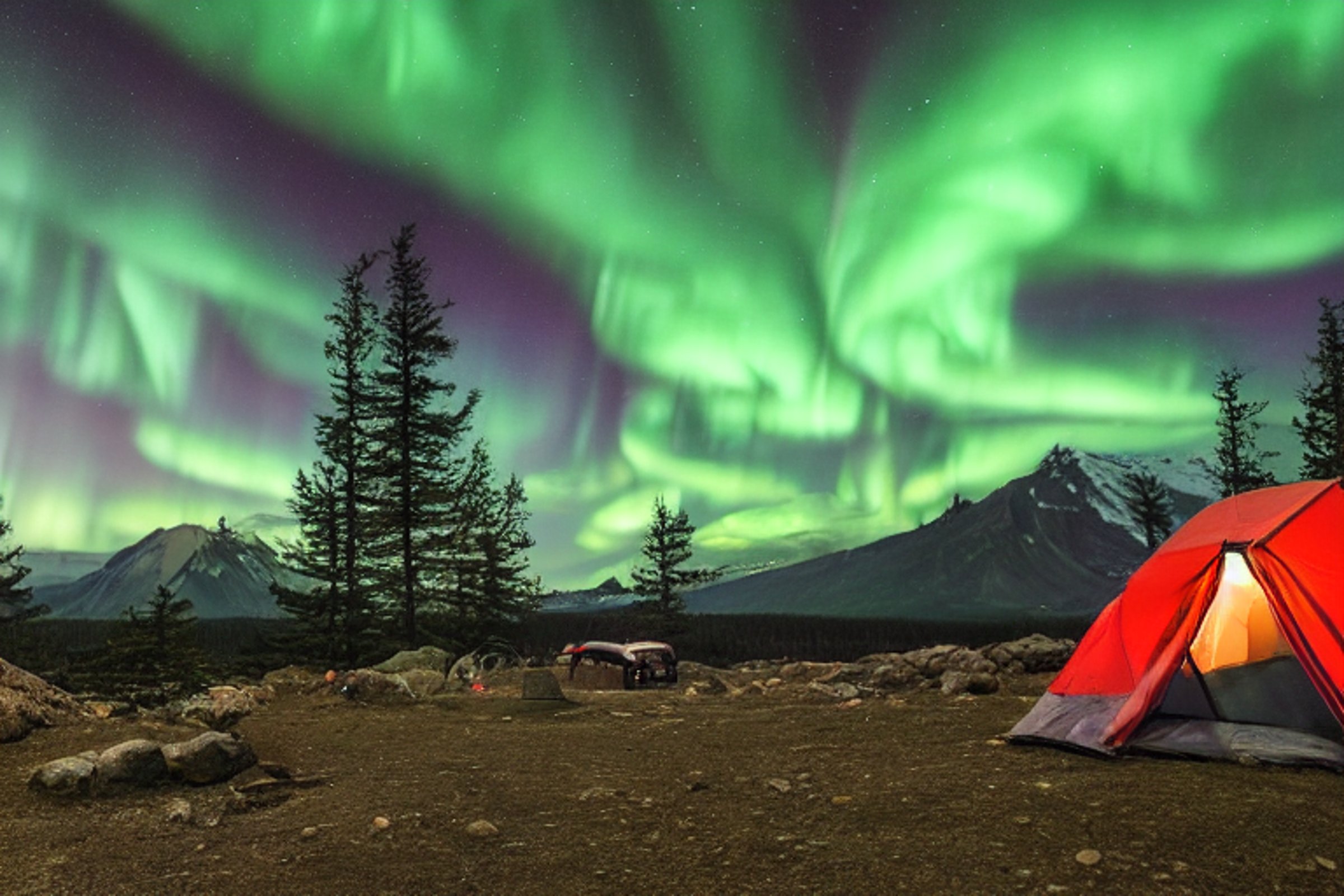 A green tent rests on a snowy mountain campsite beneath glowing northern lights. Tall trees, camping chairs, and a nearby parking spot surround the dirt clearing. Dark mountains rise in the distance under the starry sky.