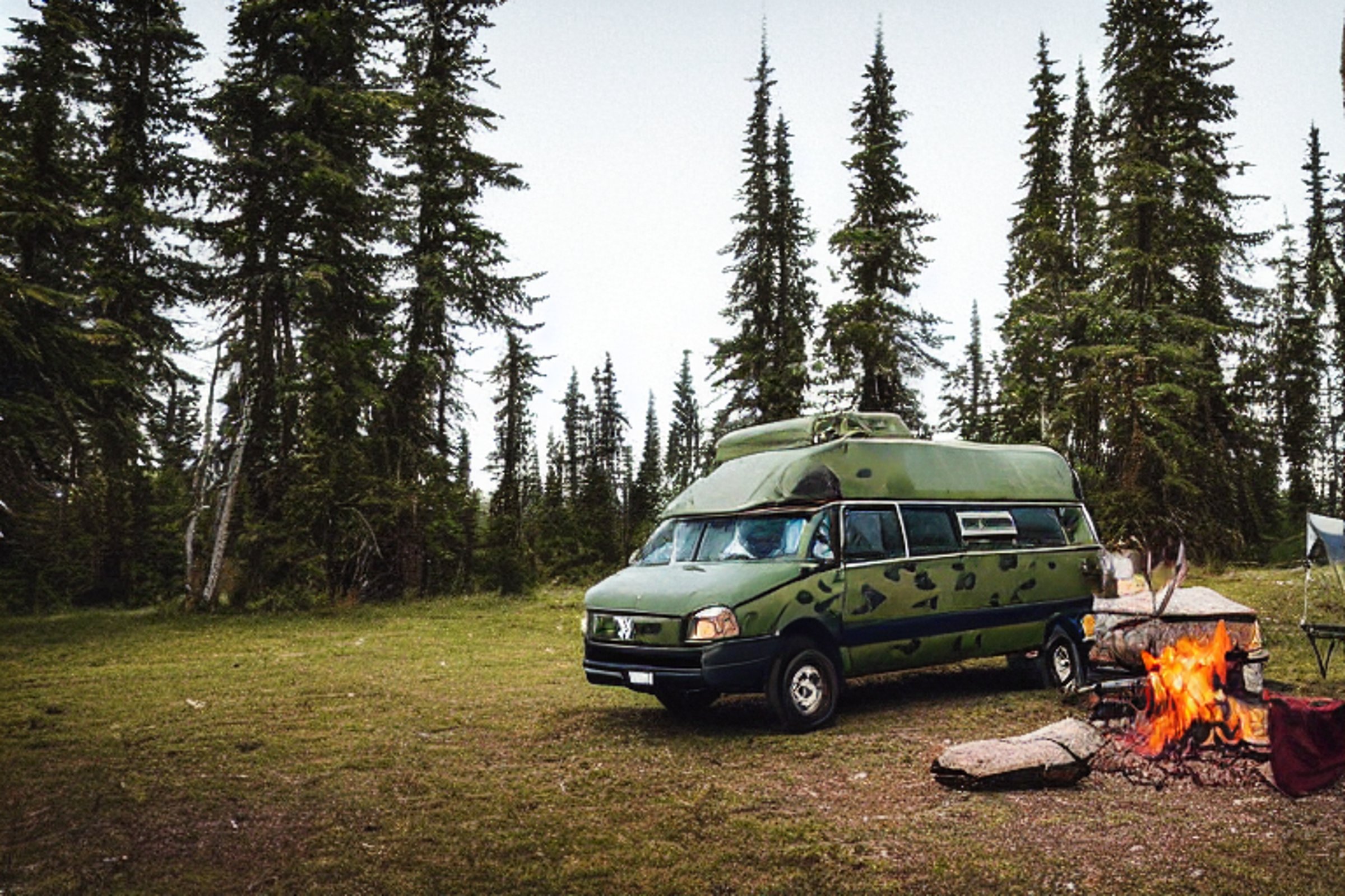 A green camo van rests on a mountain campsite beneath glowing northern lights. A warm campfire lights the chairs, logs, and guitar on the dirt. Small trees surround this peaceful night retreat.