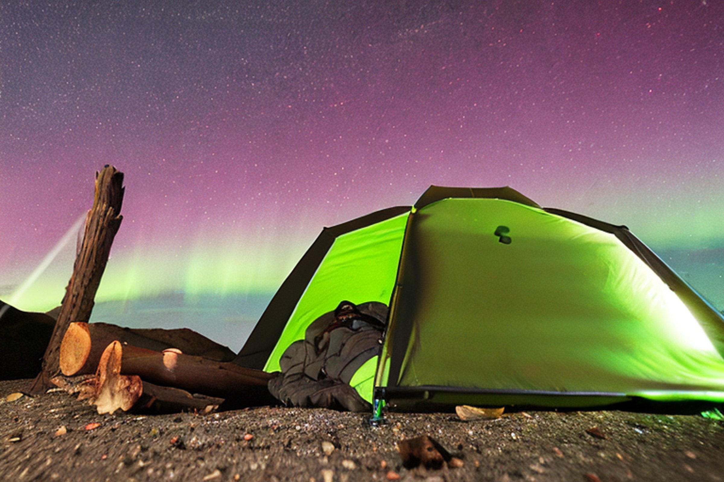 A green camo tent rests on a quiet gravel campsite beneath glowing northern lights. A warm campfire flickers nearby, with tall mountains rising in the distance. A soft light shines from inside the tent, making the dark night feel cozy.