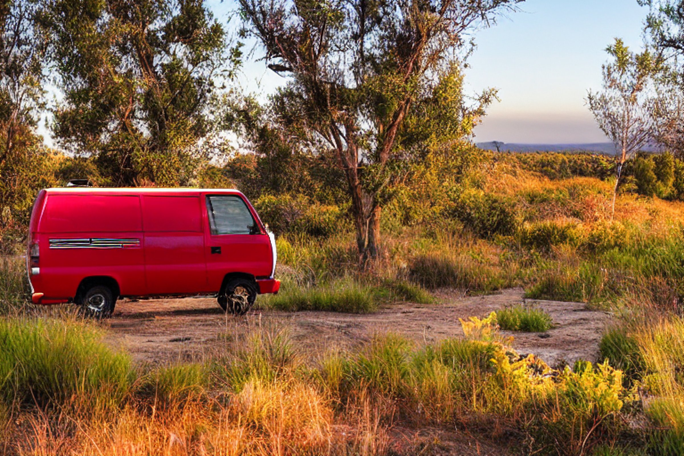 Two red camper vans rest at a sunny beach campsite in San Diego. Small streams wind through the dirt, while surfers enjoy the waves nearby. Fall-colored trees and distant mountains make the sunrise scene feel peaceful and bright.
