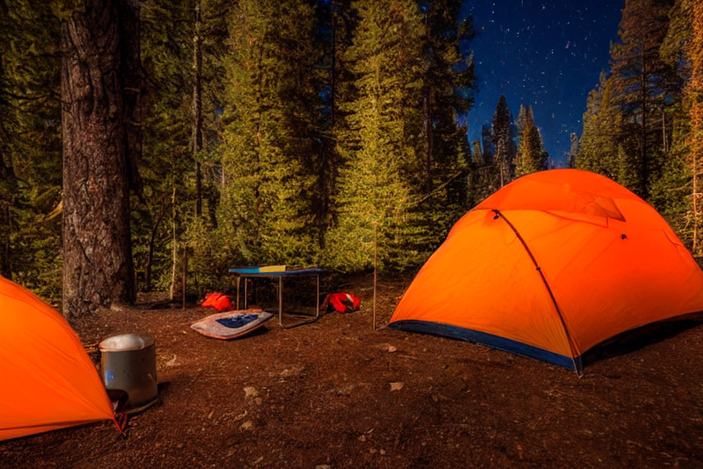 An orange tent glows beside a warm campfire under a sky full of stars. Surrounded by trees and rocky hills in Yosemite, the cozy campsite has a picnic table and a small lantern resting on the ground.