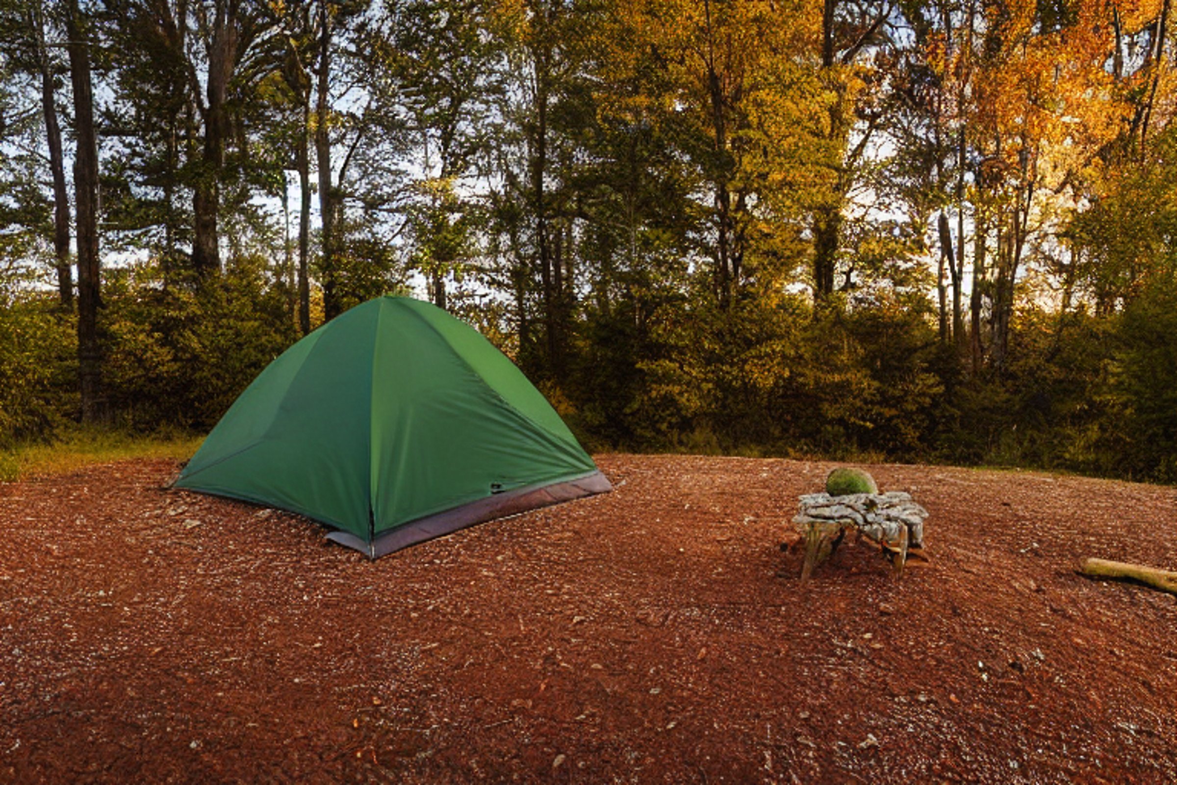 A green and tan tent rests beside a warm campfire near a quiet lake. Golden light shines through colorful fall trees, while rocky ground and berry-covered bushes frame the campsite. A pale moon hangs in the clear sky.