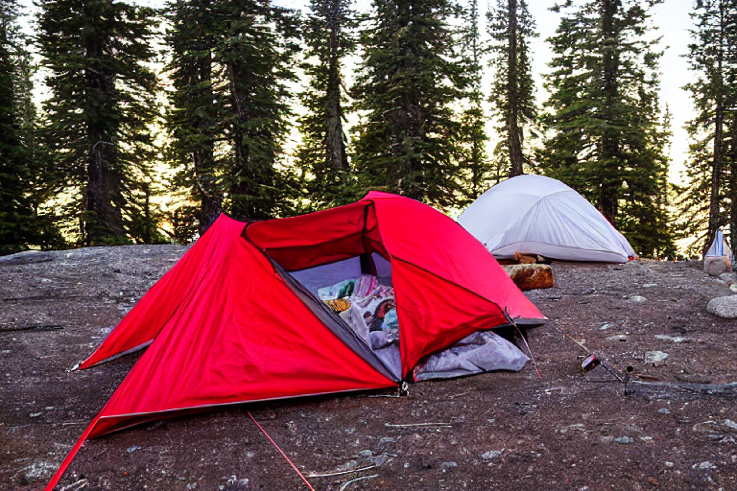 A red and white tent rests on a snowy campsite as a warm campfire glows nearby. Clothes hang from a simple line, with camping gear on the ground. Tall mountains rise in the distance beneath the soft sunset sky.