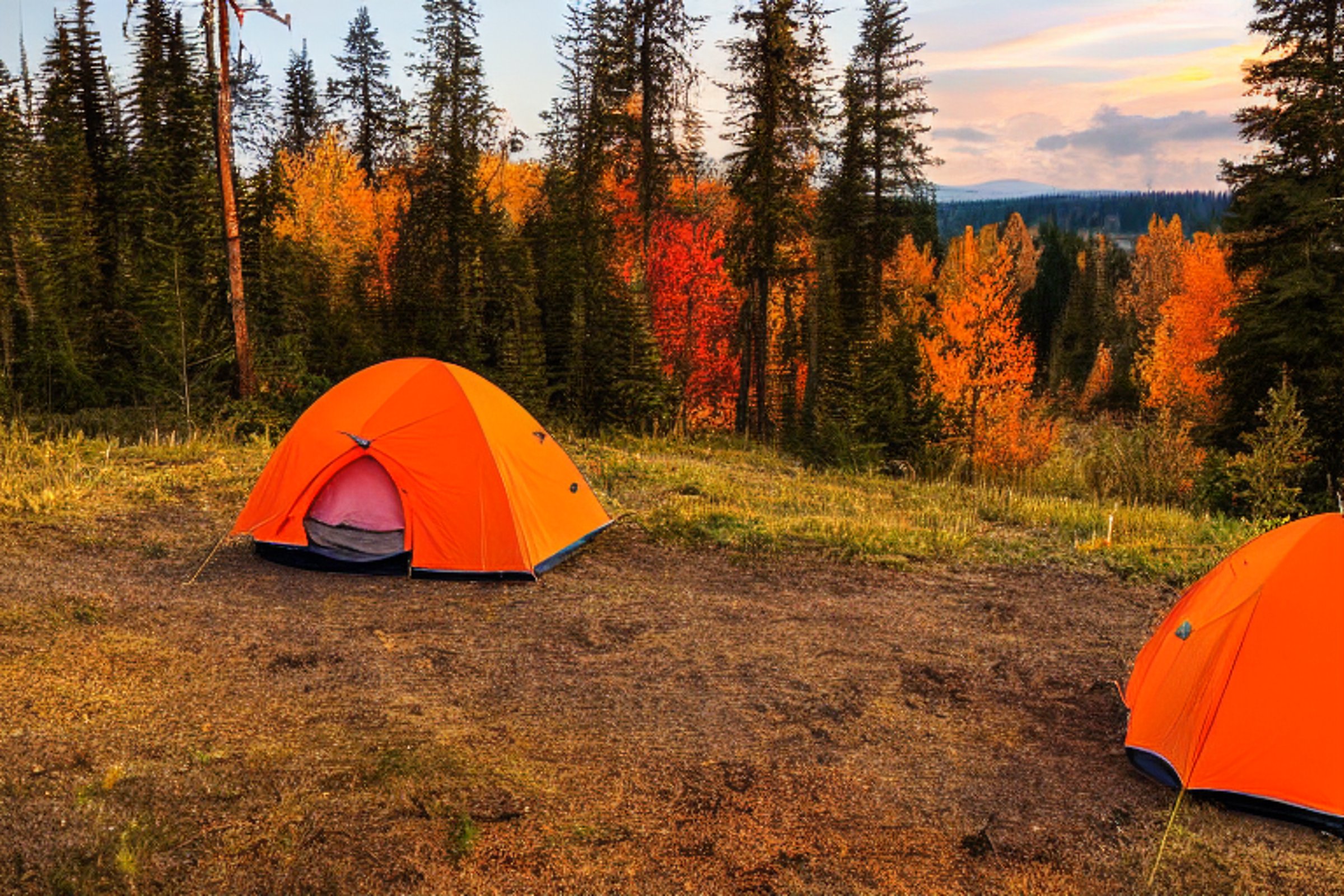 An orange tent rests beside a warm campfire on a sunny dirt clearing. Fall-colored trees, tall forests, and rolling hills surround the campsite. Two camping chairs invite you to sit and enjoy the peaceful sunset.