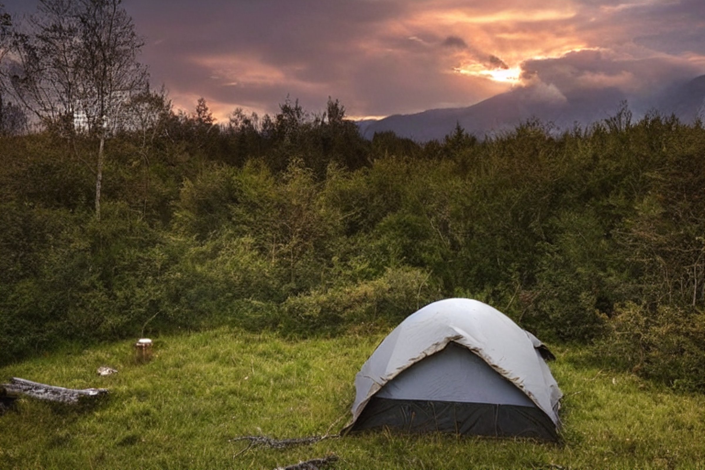 A brown tent rests on a mountain campsite as rain falls beneath a glowing sunset. Overgrown grass, small trees, and gravel patches surround a stone-ringed campfire and scattered logs.