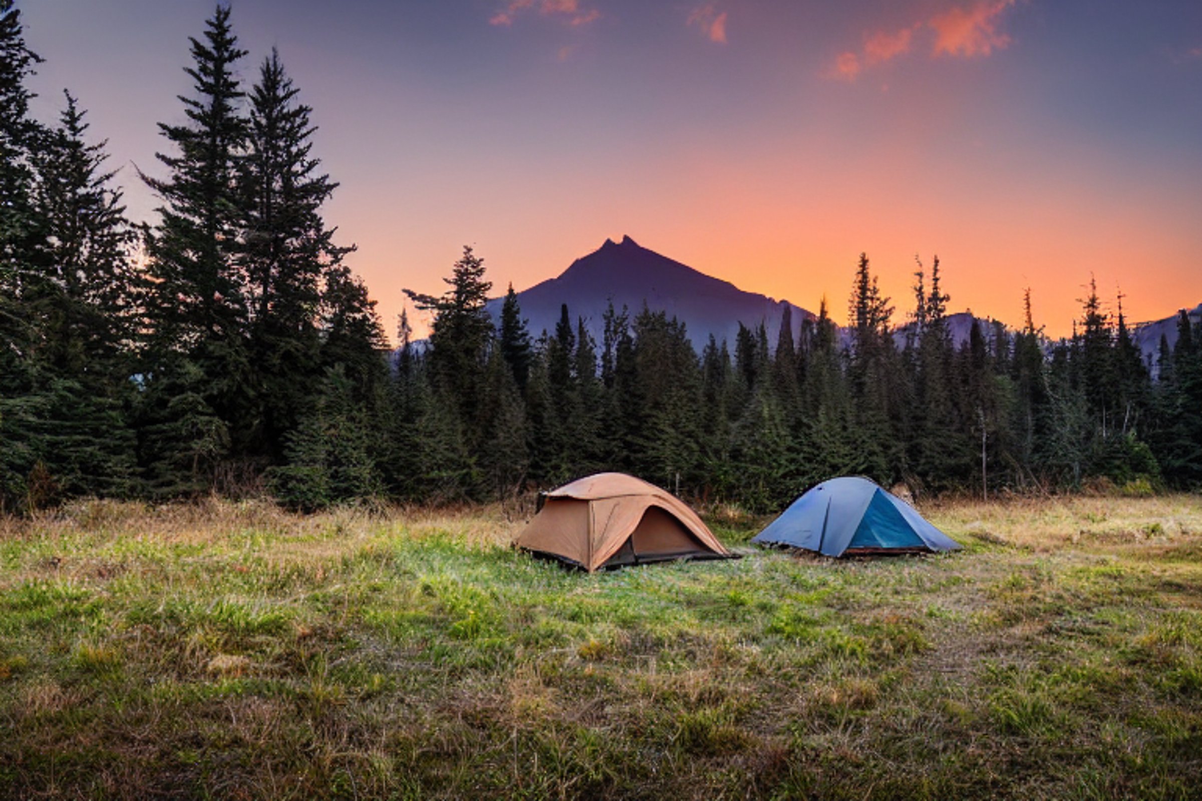 A cozy brown tent rests beside a warm campfire on Vancouver Island. A dirt path winds through tall grass toward rolling hills and distant mountains, glowing under a clear sunset sky.