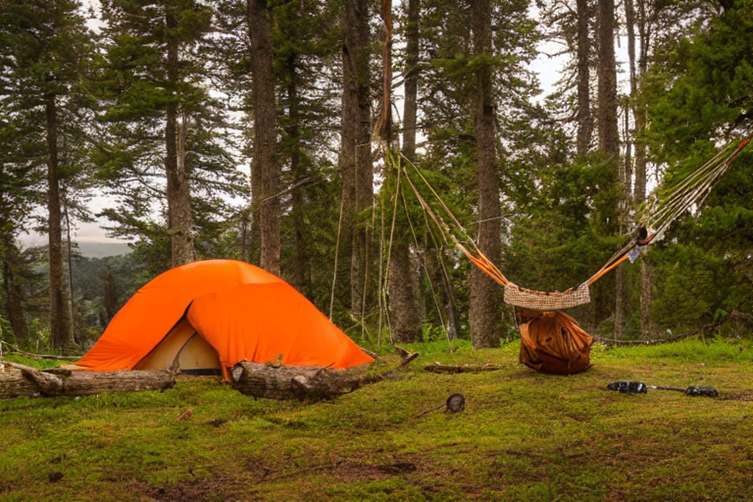 An orange tent rests beside a warm hot spring as the sun sets under stormy skies. A campfire glows near a large log, while a hammock hangs between the trees. Fallen leaves and rough ground give the campsite a wild, cozy feel.