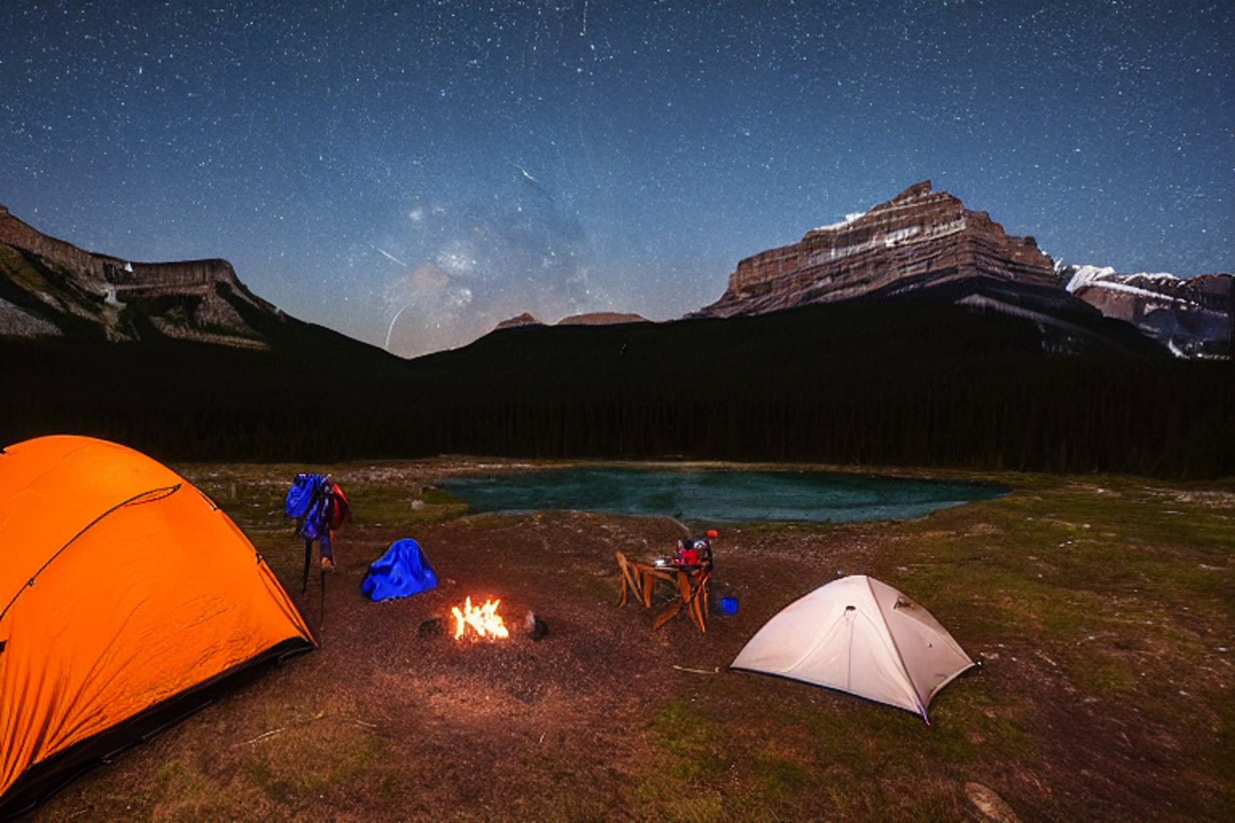 A cozy brown tent rests beside a glowing campfire in a foggy mountain valley. Stars shine above the dark peaks, while a backpack and scattered twigs sit on the leafy ground.