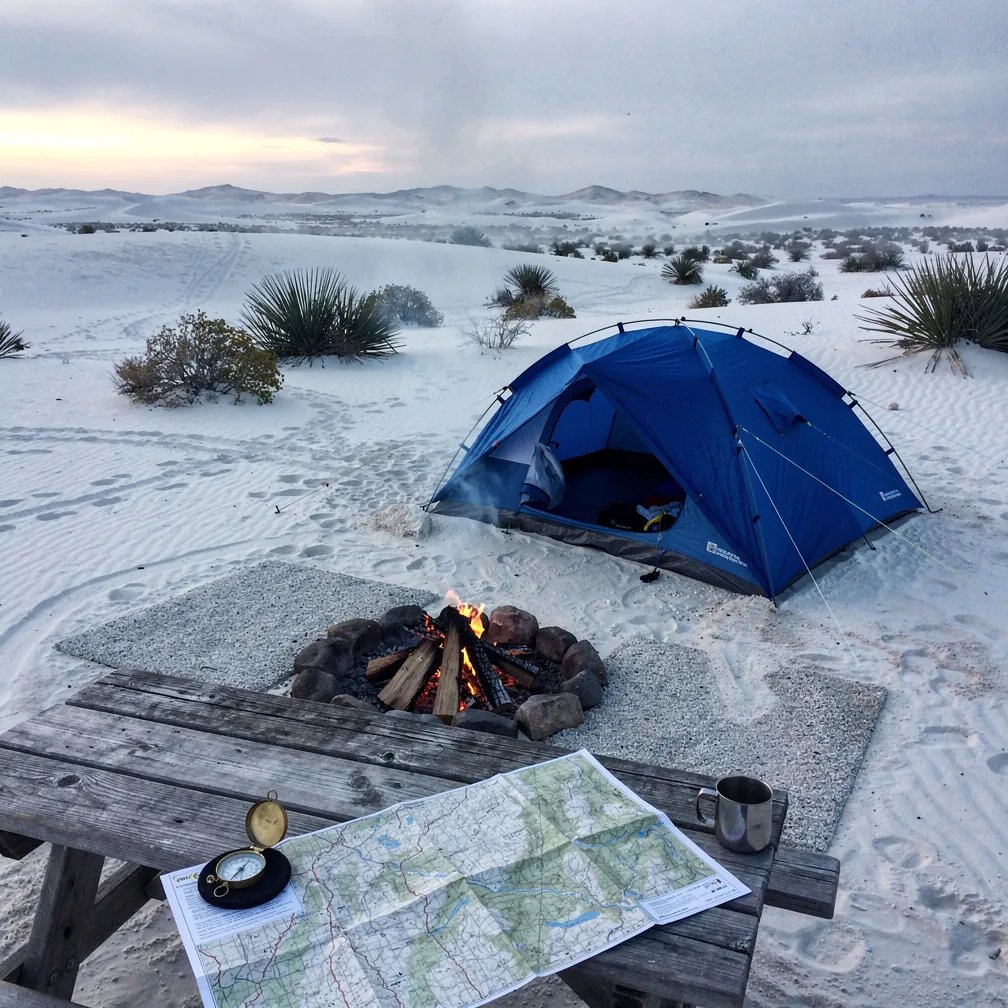 A blue tent sits in smooth white sand dunes as the sky wakes up after a storm. A small fire ring glows with warm embers and a thin wisp of smoke drifts into the cool air. Nearby, a weathered picnic table holds a map and compass, ready for a quiet morning adventure.
