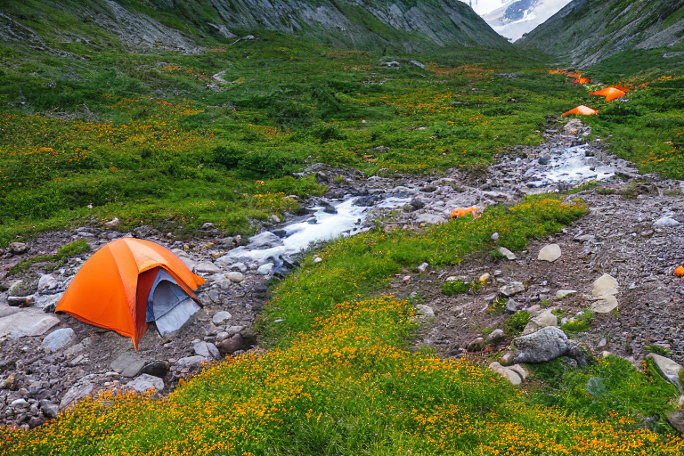 An orange tent glows warmly beside a clear stream in a rocky campsite. Golden sunlight lights the valley, bushes, and colorful mountain scenery around it.