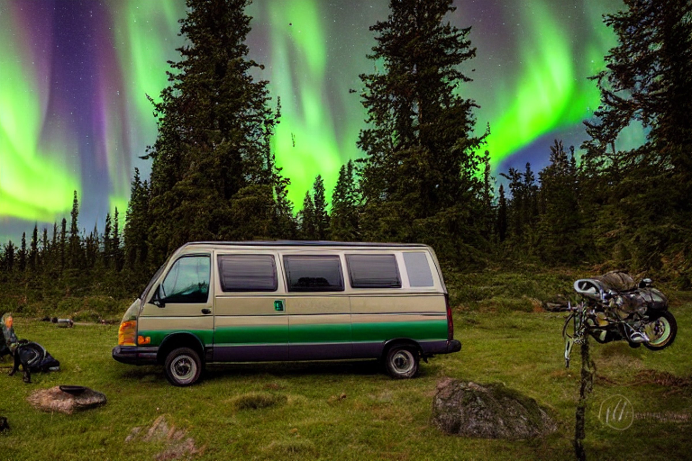 A forest-green camper van rests on a rough dirt clearing beneath glowing northern lights. Small trees surround the campsite, creating a quiet, wild feeling. Nearby surfers add a fun touch to this peaceful Washington night.