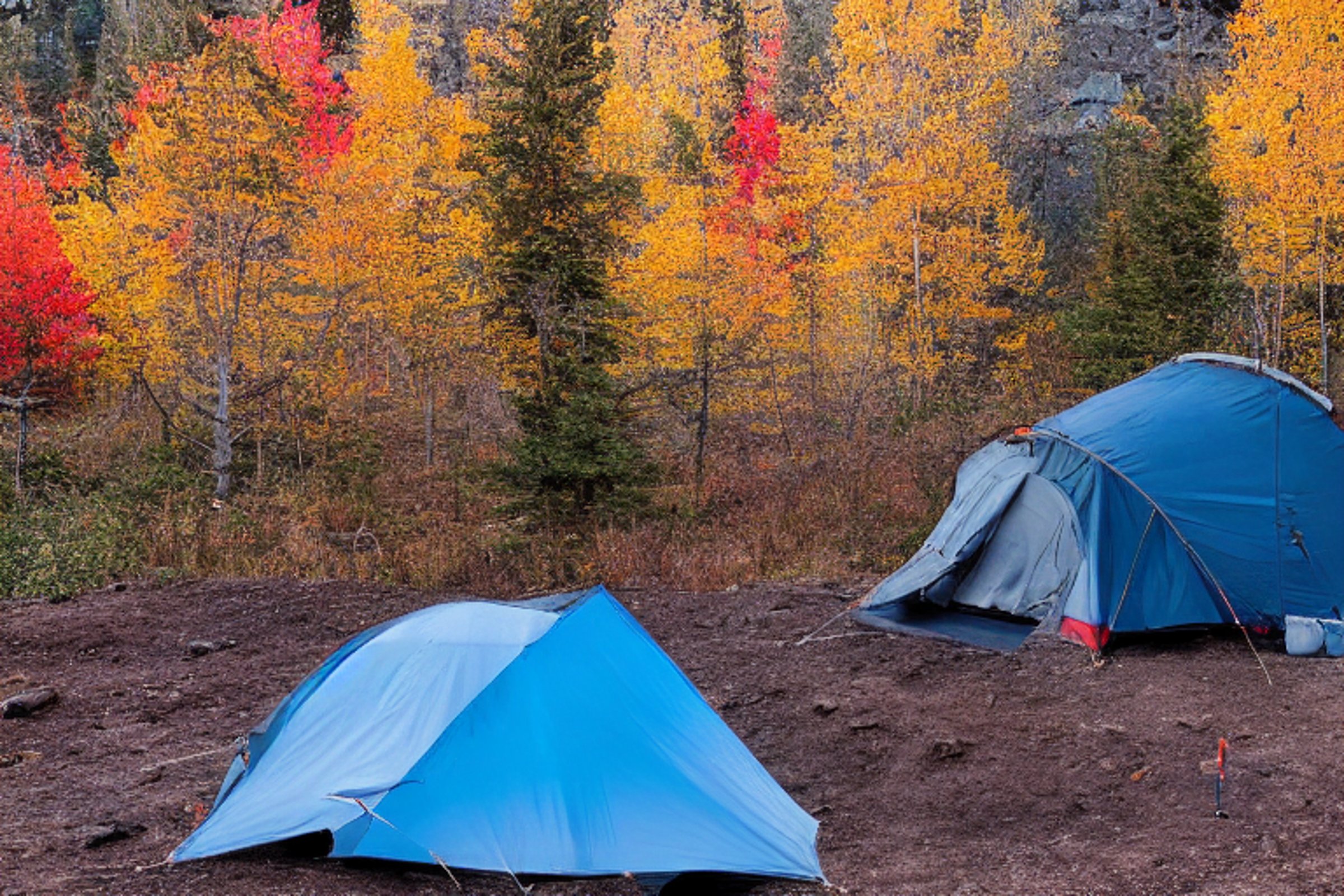 A black tent rests on a quiet mountain campsite as dusk settles in. Colorful fall trees surround the dirt clearing, with camping chairs and a backpack nearby. The clear sky and cool evening light create a peaceful outdoor escape.