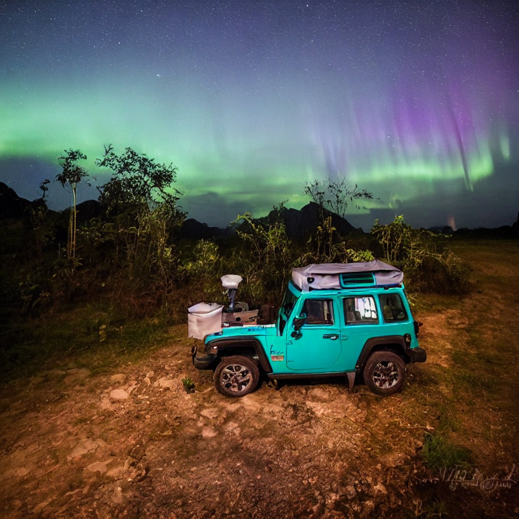 A teal Jeep with an open rooftop tent rests on a quiet dirt campsite in Halong Bay. Small trees and tall grass surround the camp, while a warm lantern glows nearby. Above, bright green auroras ripple across the clear night sky.