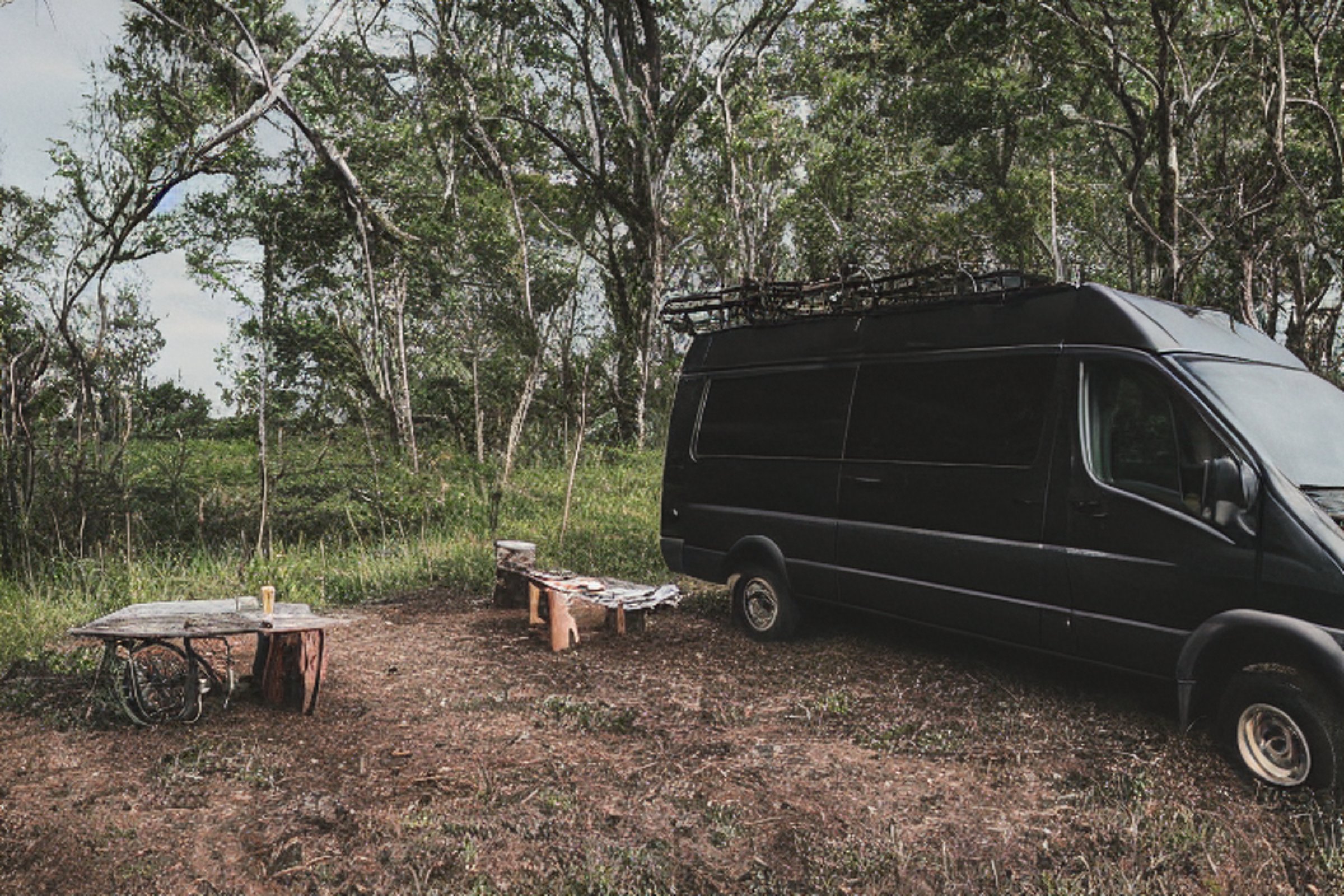 A black camper van rests on a quiet dirt campsite beneath a clear dusk sky. Large stone slabs and a winding hiking path surround the camp, while clothes sway on a nearby line. A deer stands far away in the peaceful landscape.