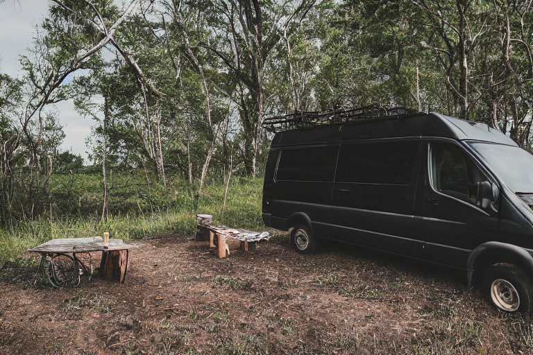 A black camper van rests on a quiet dirt campsite beneath a clear dusk sky. Large stone slabs and a winding hiking path surround the camp, while clothes sway on a nearby line. A deer stands far away in the peaceful landscape.