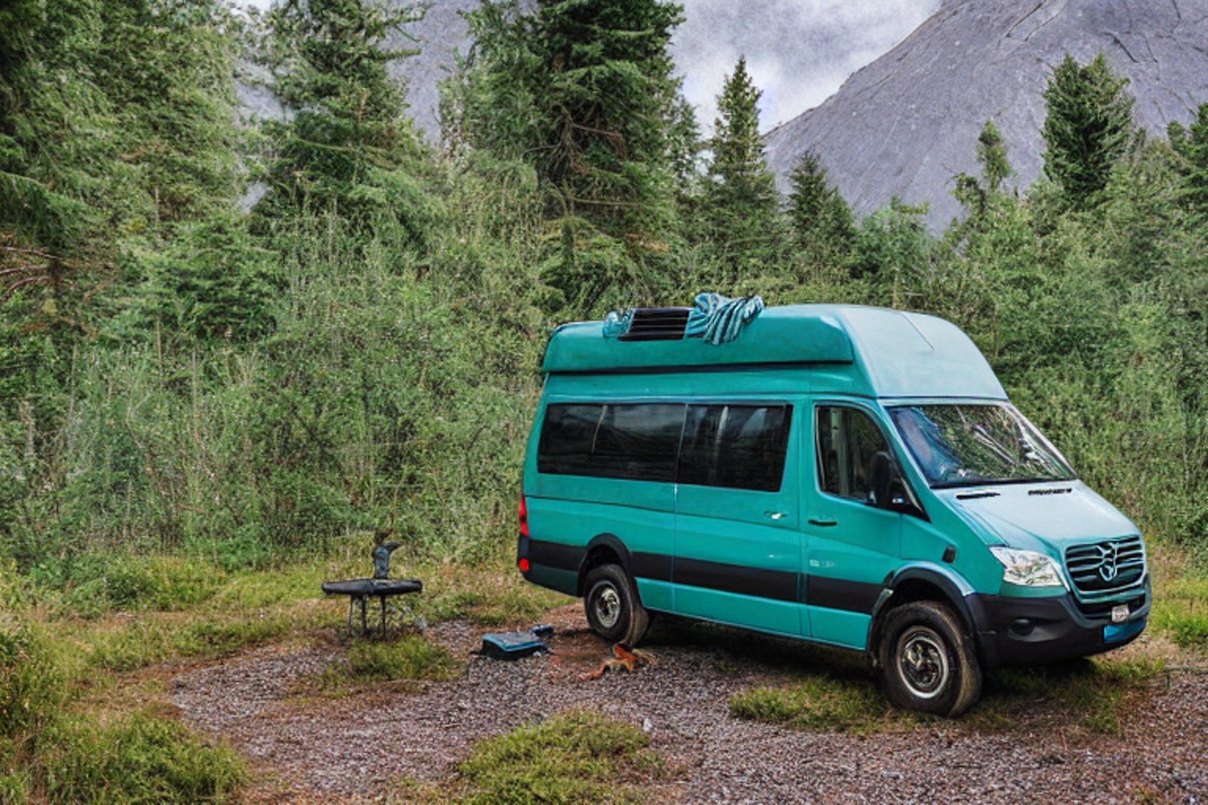 A teal camper van rests on a quiet dirt patch in a foggy forest. Large bushes, scattered stones, and a rocky foreground surround the campsite, while a shadowy mountain rises in the distance.