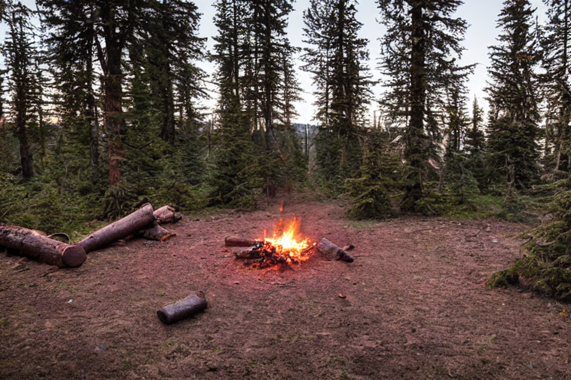 A brown tent rests on a rough, snowy campsite in Washington State. A warm campfire glows among the logs and boulders as the sun sets over the rugged land.