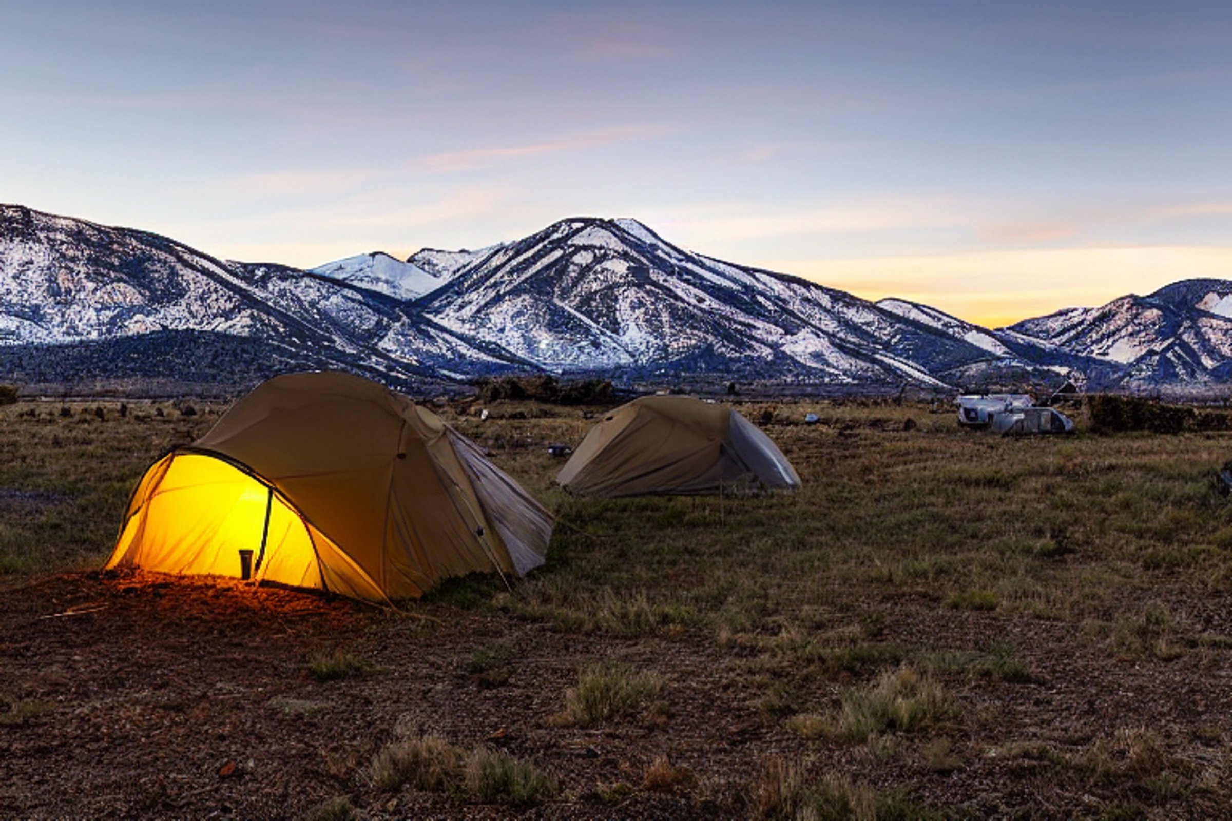 A cozy tent rests on a dirt patch at a paved campsite beneath a snowy sunset sky. Mountains rise in the distance, while a deer wanders across the Idaho plains. A glowing lantern adds warmth to the quiet scene.