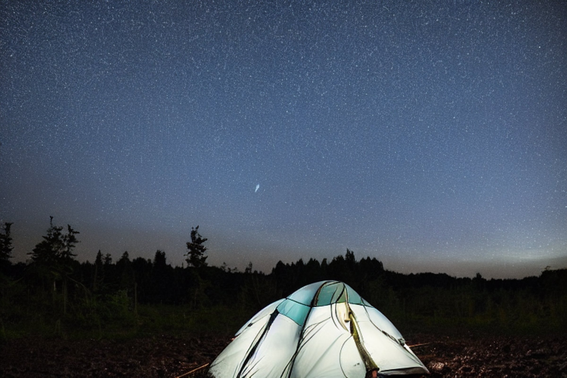A bright blue tent rests on a dirt campsite beside a quiet lake. Rainy air surrounds the camp, while stars shine above and stones scatter across the ground. A warm lantern glows nearby, and a person’s flashlight lights the night in the distance.
