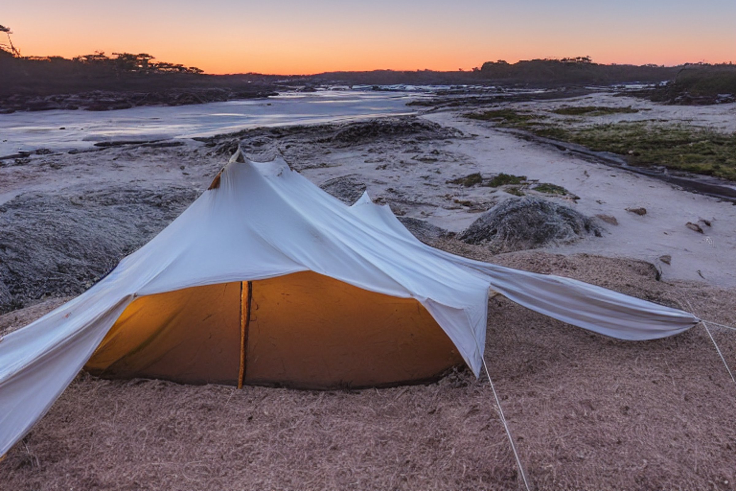 A tan tent rests on a sandy beach beneath a sky full of bright stars. Boulders, a dirt campsite, and a small parking spot sit near the snowy ocean, while clothes sway on a nearby line.
