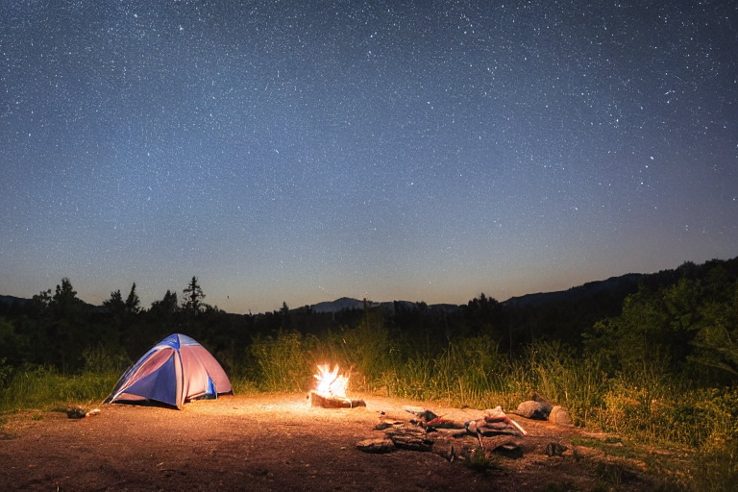 A blue tent glows beside a warm campfire under a cloudy, starry sky. Set near a hot spring, the rough campsite has large rocks, camping chairs, and a clothesline swaying in the night air.