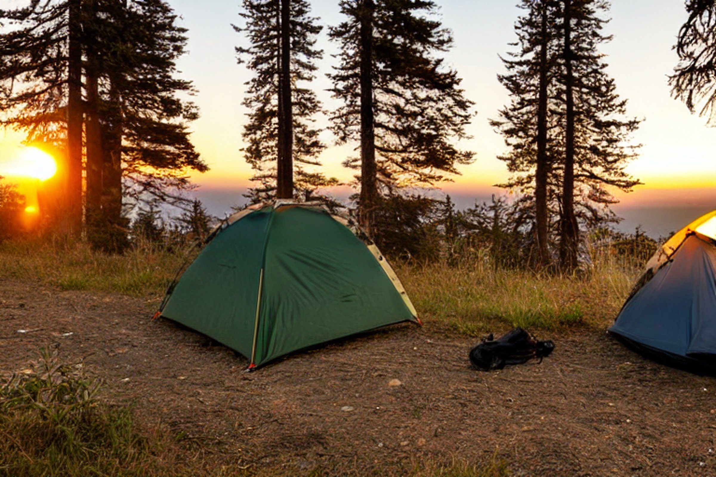 A forest-green tent rests beside a warm campfire on a quiet patch of earth along the California coast. Rolling hills and a clear sunset sky stretch into the distance, with the moon rising above. A backpack and clothesline add a cozy, lived-in touch.