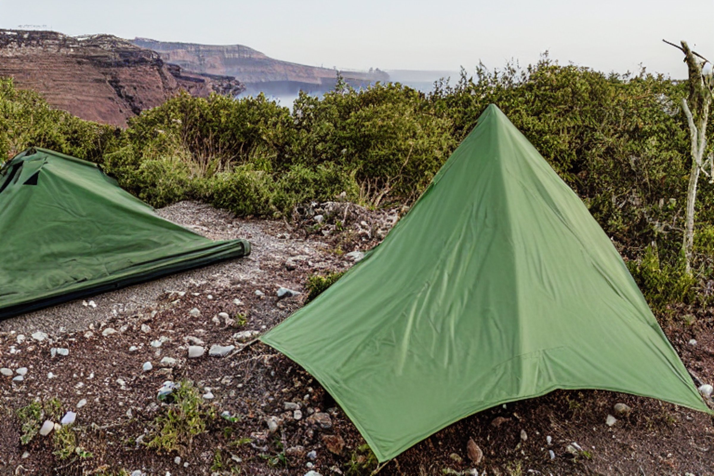 A green camo tent rests on a paved campsite dusted with snow in Santorini. A picnic table, stone gravel, and a winding dirt path create a quiet outdoor retreat.