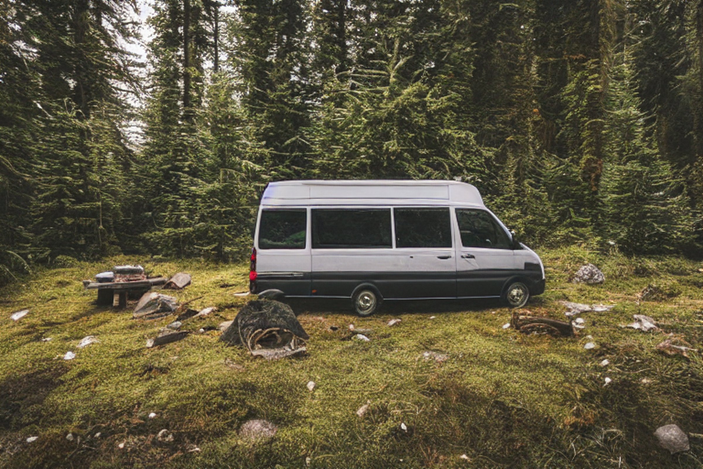 A black camper van rests beside a warm campfire in a snowy mountain valley. Mossy trees, scattered stones, and a large log surround the dirt campsite, while sunset colors glow behind the peaks.