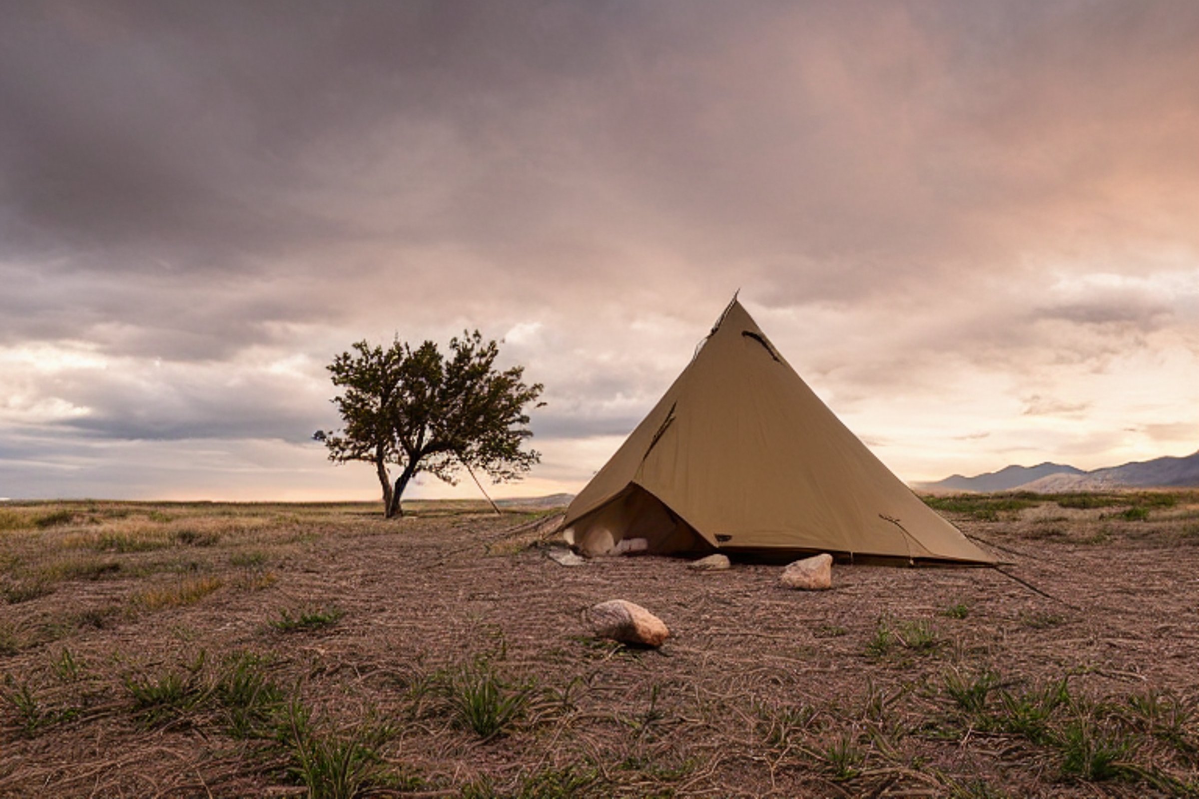A green and tan tent rests on a paved campsite at dusk in Great Sand Dunes National Park. Large stone slabs, fallen leaves, and a small twig surround the dirt patch. A hammock hangs between the trees under a cloudy evening sky.