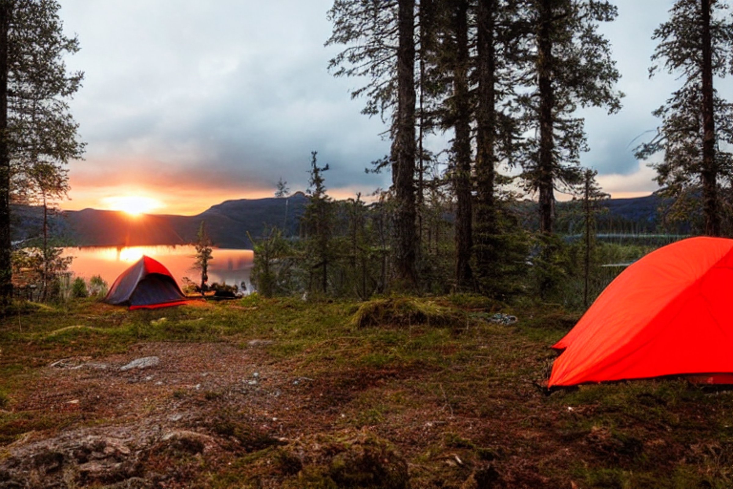 A bright red tent rests on a quiet dirt clearing in the Norwegian wilderness. A warm campfire glows beside a large log, while the tent shines softly from within. Camping gear and a backpack sit nearby under the cloudy sunset sky.