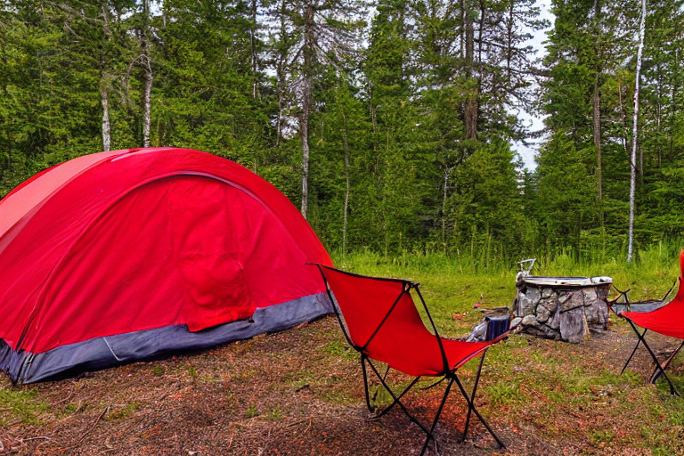 A red tent glows beside a warm campfire under a clear night sky. Camping chairs sit on a leafy, gravel-strewn clearing, with dark Maine mountains rising in the distance.