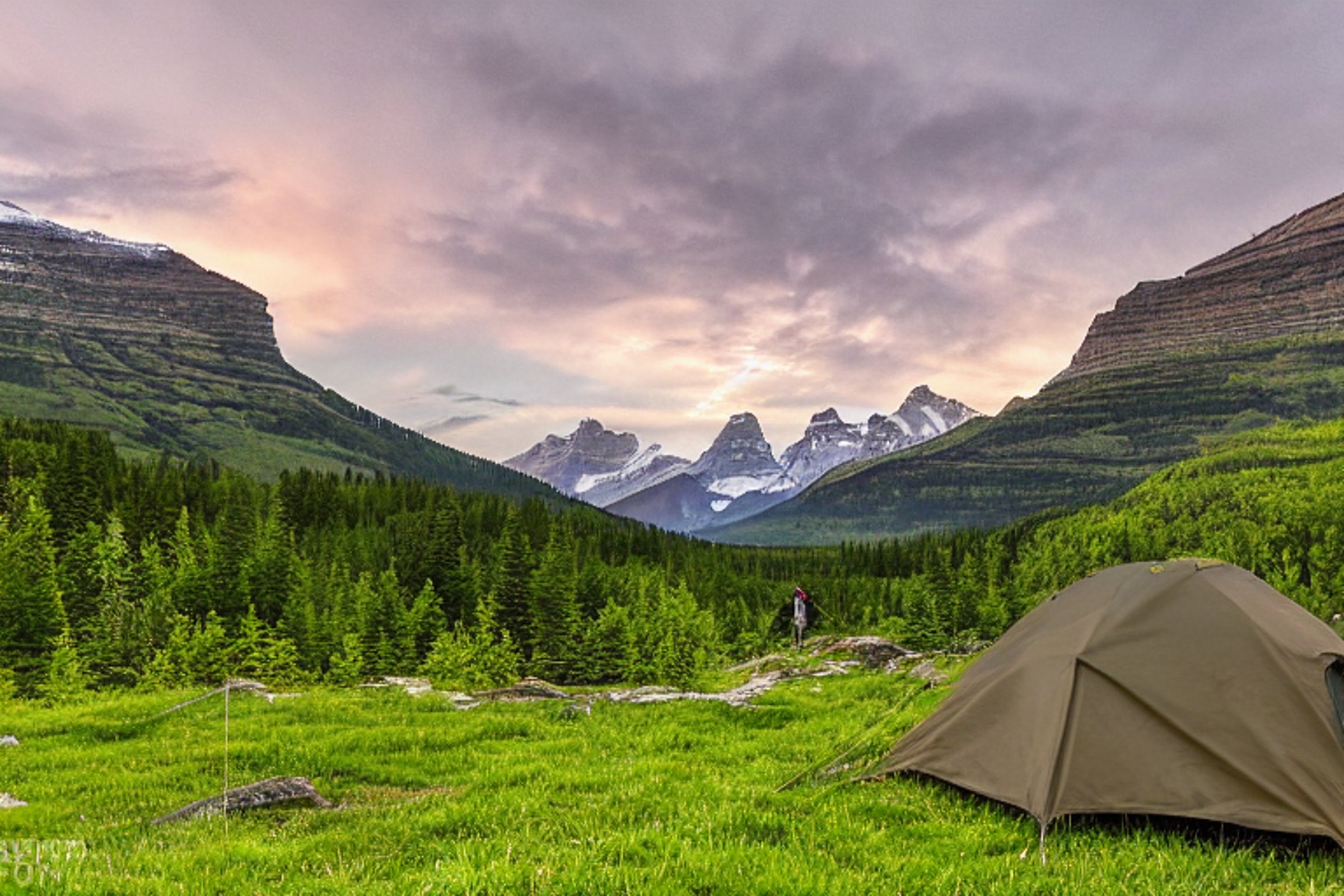 A green and tan tent rests on a quiet dirt campsite beneath a star-filled sky. Rolling hills and large stone slabs surround the camp, while a distant flashlight glows in the night.