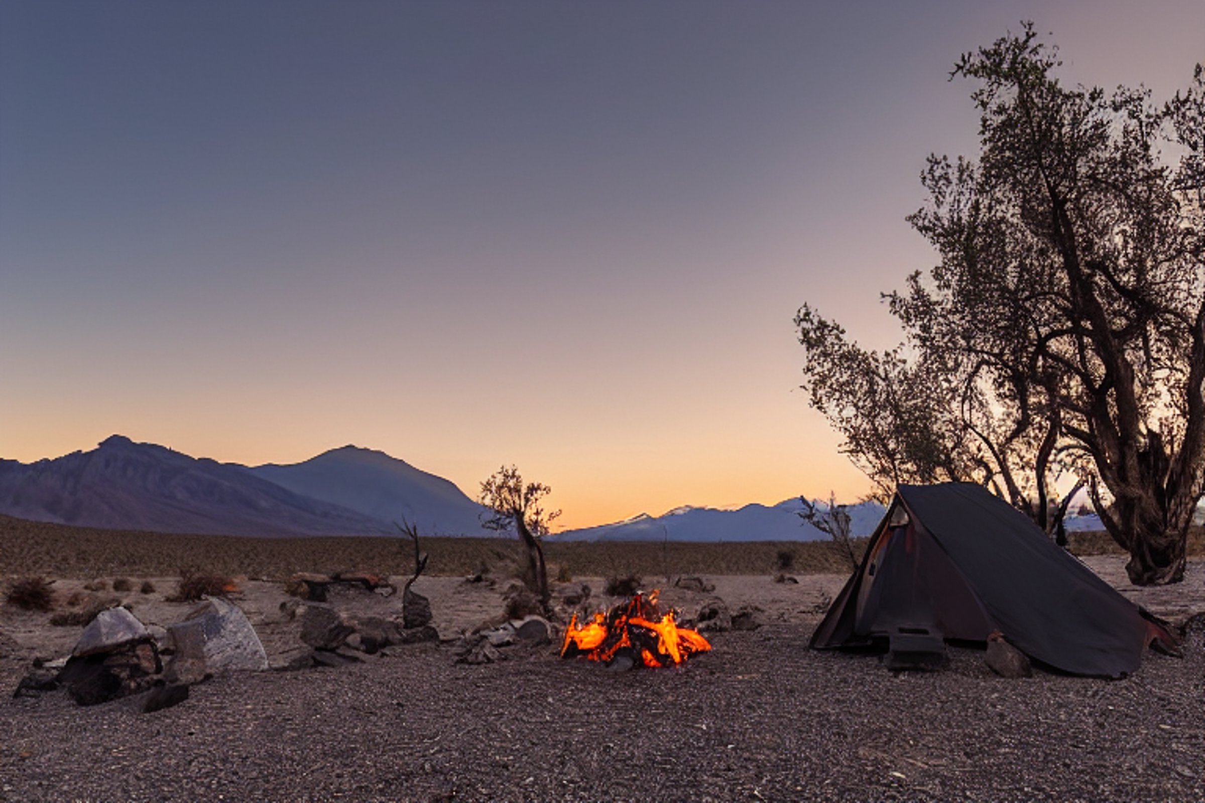 A black tent rests on a dirt campsite as the sun sets behind the mountains of Death Valley. A warm campfire glows among a ring of stones, with logs and a wooden guitar nearby. Hikers enjoy the quiet, sunny evening.