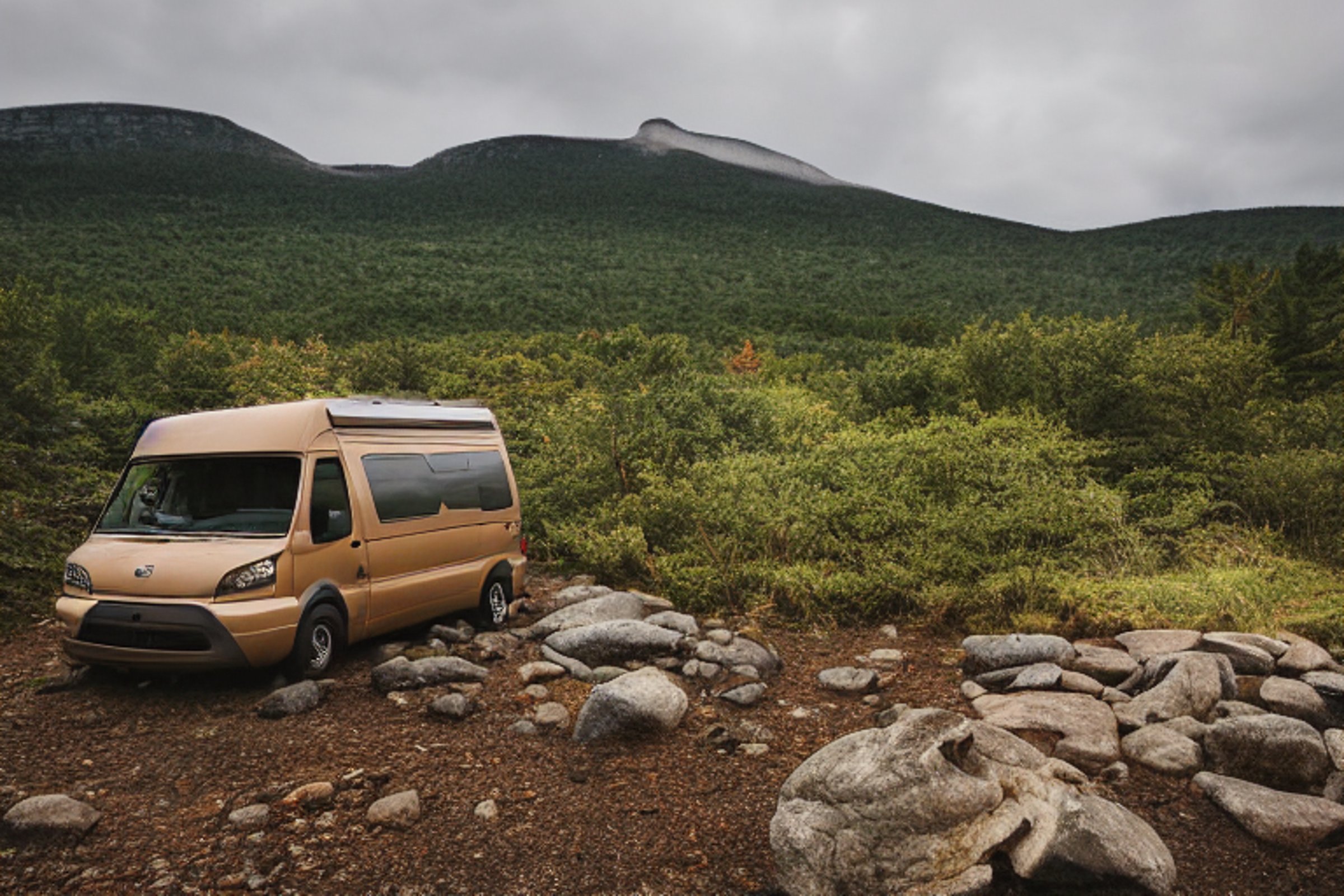 A brown camper van rests at a quiet campsite in Acadia National Park. Stone slabs and boulders surround the gravel ground, while hikers gather nearby. Above, stars peek through the stormy night sky.