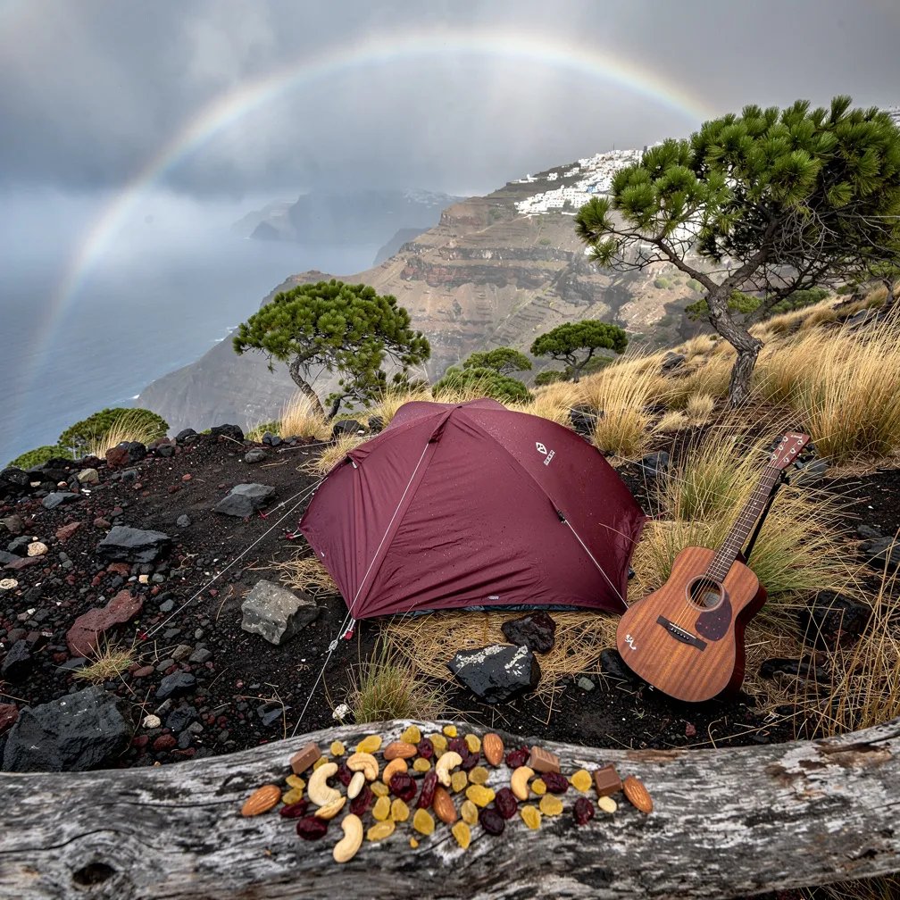 A small burgundy tent sits on a windy hillside above dark, rocky land, with tall dry grass and a few pine trees all around. Mist drifts through the trees, and a pale rainbow in the fog hangs in the distance. An acoustic guitar rests by the tent, and trail mix is spilled across an old log like a snack left behind. The air feels cool, quiet, and fresh after the rain.
