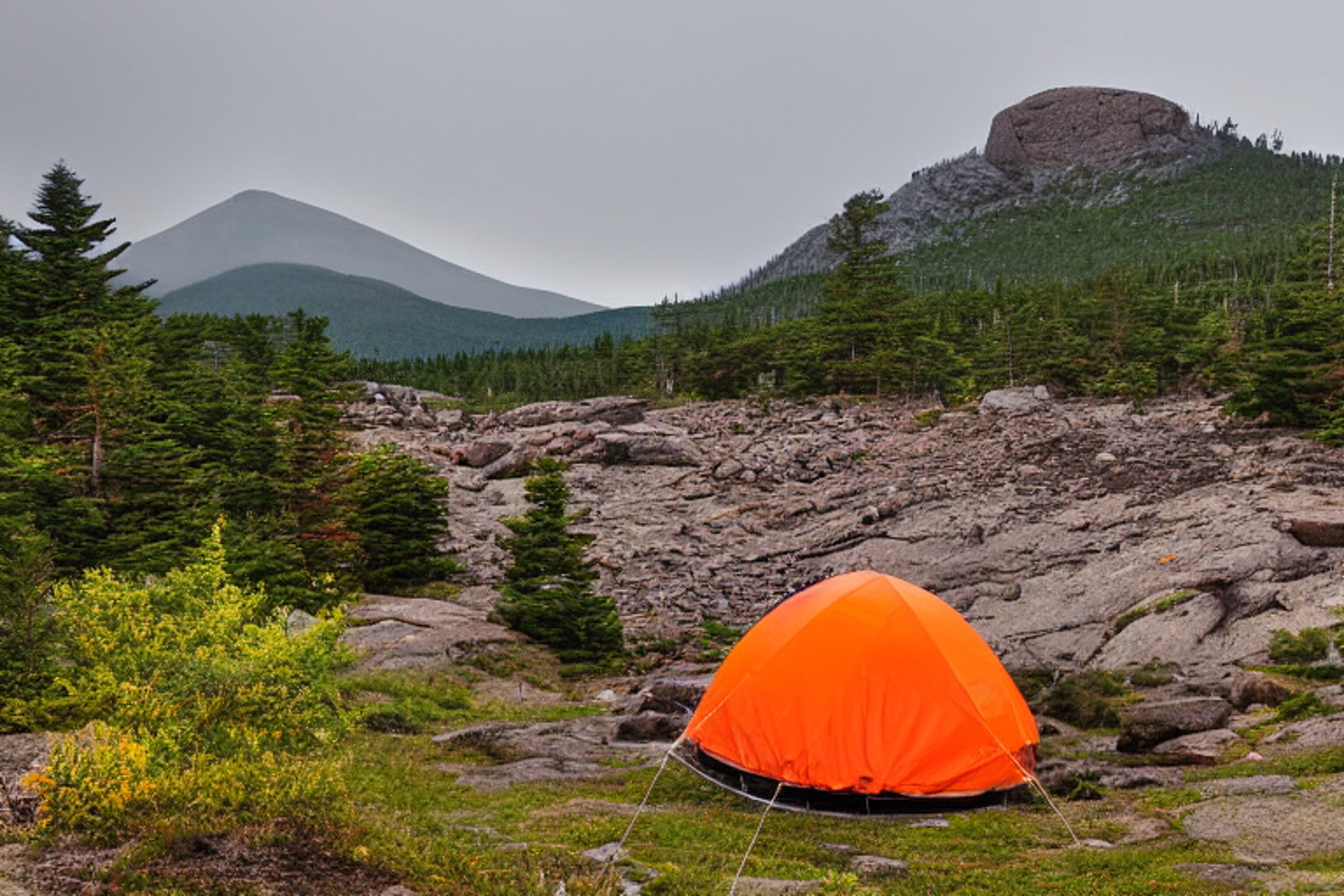 An orange tent rests on a quiet dirt campsite beneath a sky full of stars. A clear stream winds past the boulders, while misty mountains rise in the background. Clothes hang from a line, adding a cozy touch to this peaceful Acadia night.