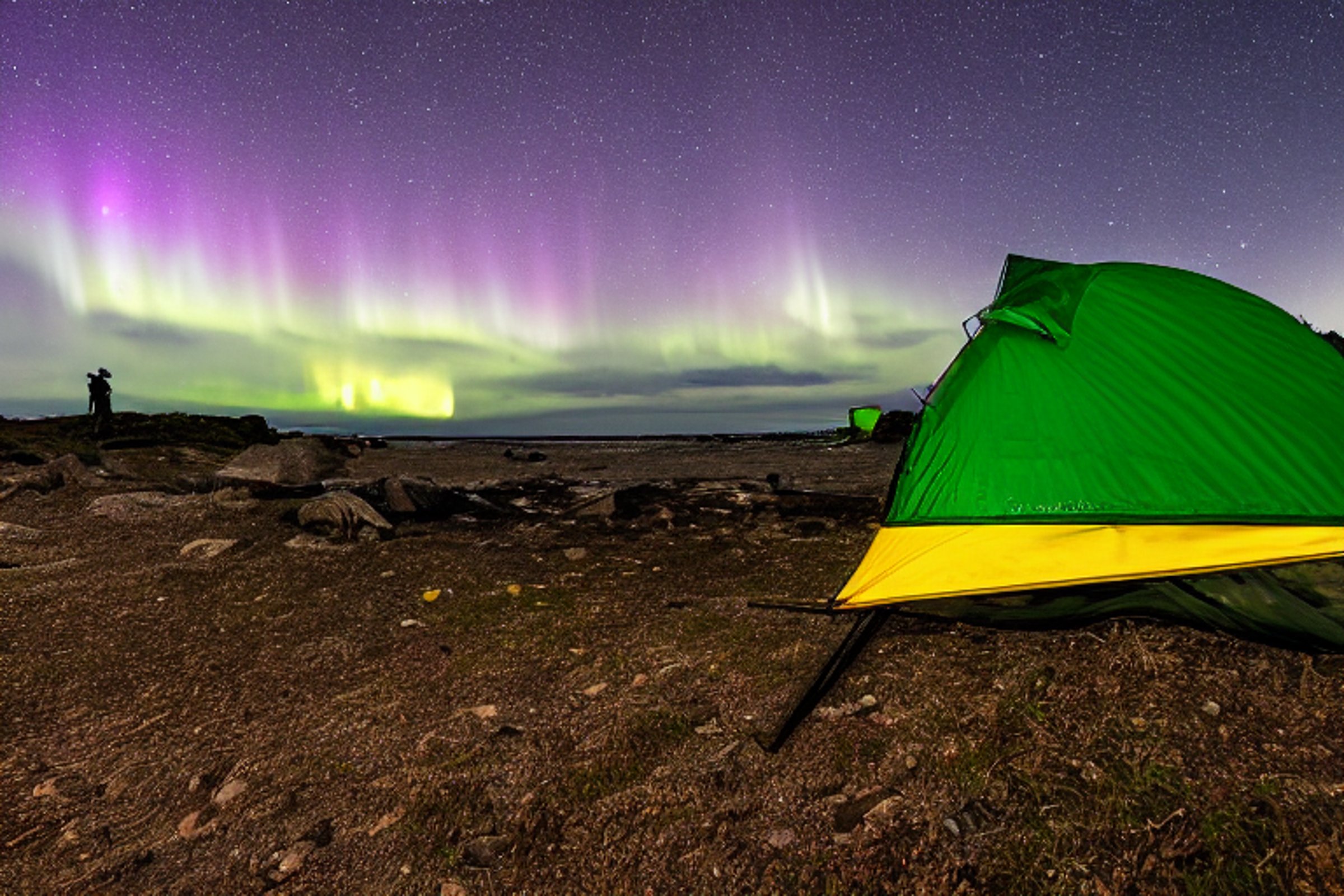 A green camo tent rests on a dirt patch beside a paved beach campsite. Waves roll nearby as hikers explore the shore, while camping gear and a backpack sit by the tent. Above, cloudy skies glow with colorful northern lights and a distant moon.