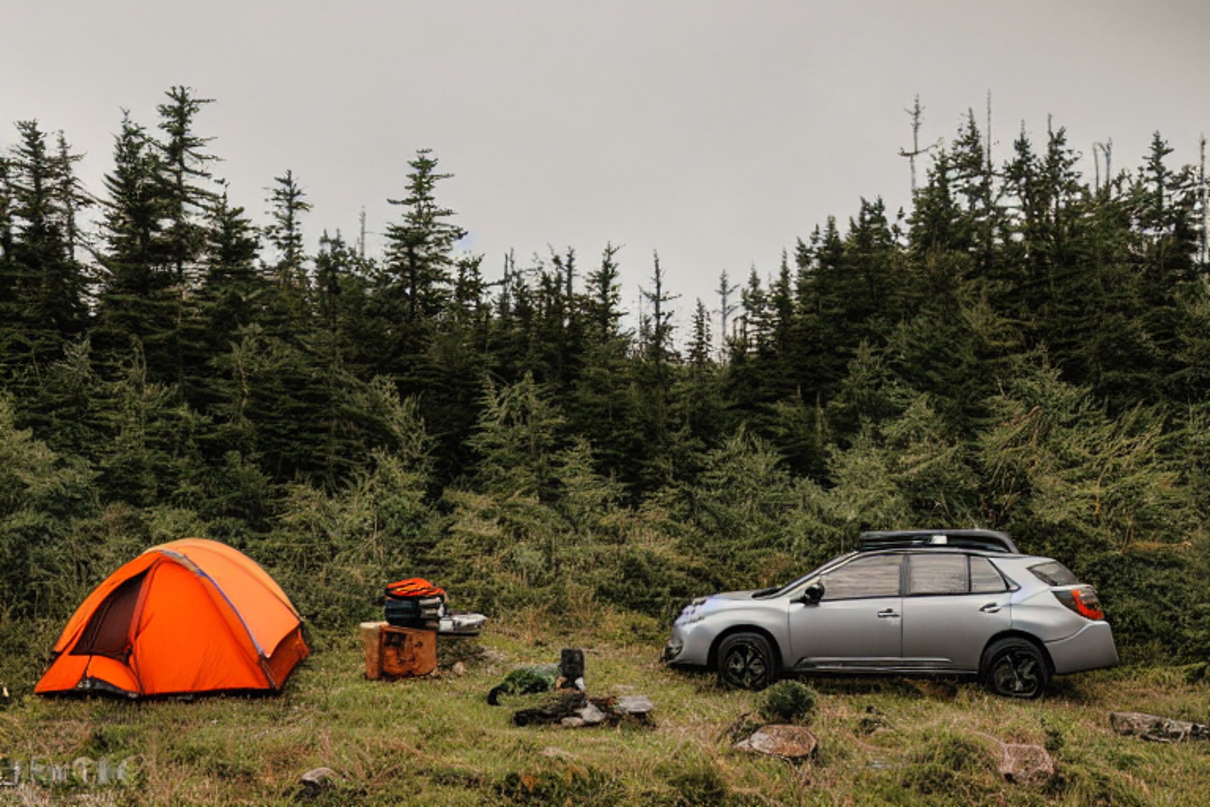 A gray and orange Subaru rests beside a cozy rooftop tent on the rainy Maine coast. Ferns and large stone slabs surround the dirt campsite, while a small campfire glows among the stones. A backpack sits nearby, ready for adventure.