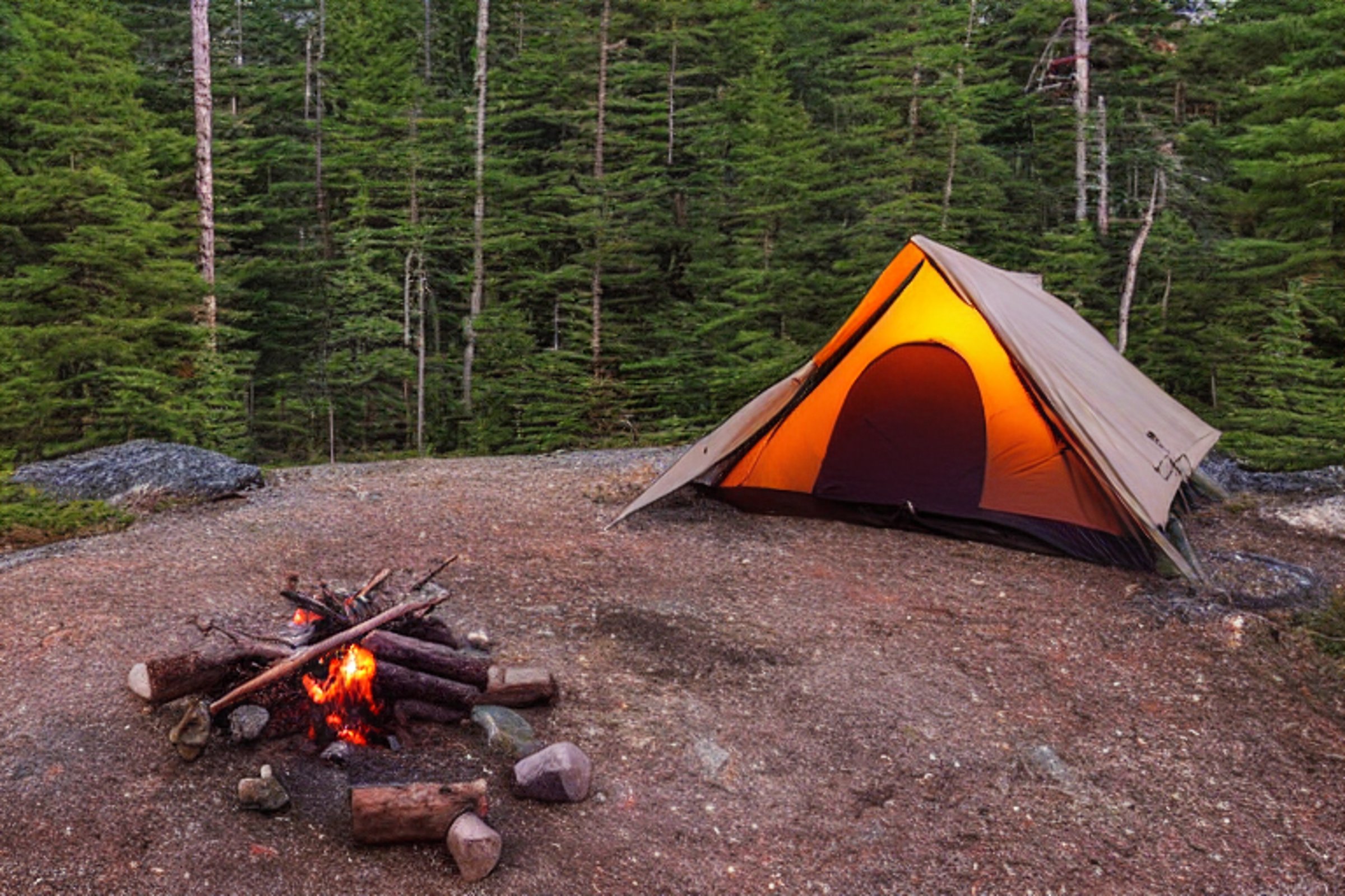 A cozy brown tent rests on a dirt clearing in Acadia National Park. A warm campfire glows among a ring of stones, while fog drifts through the dark, rocky woods.