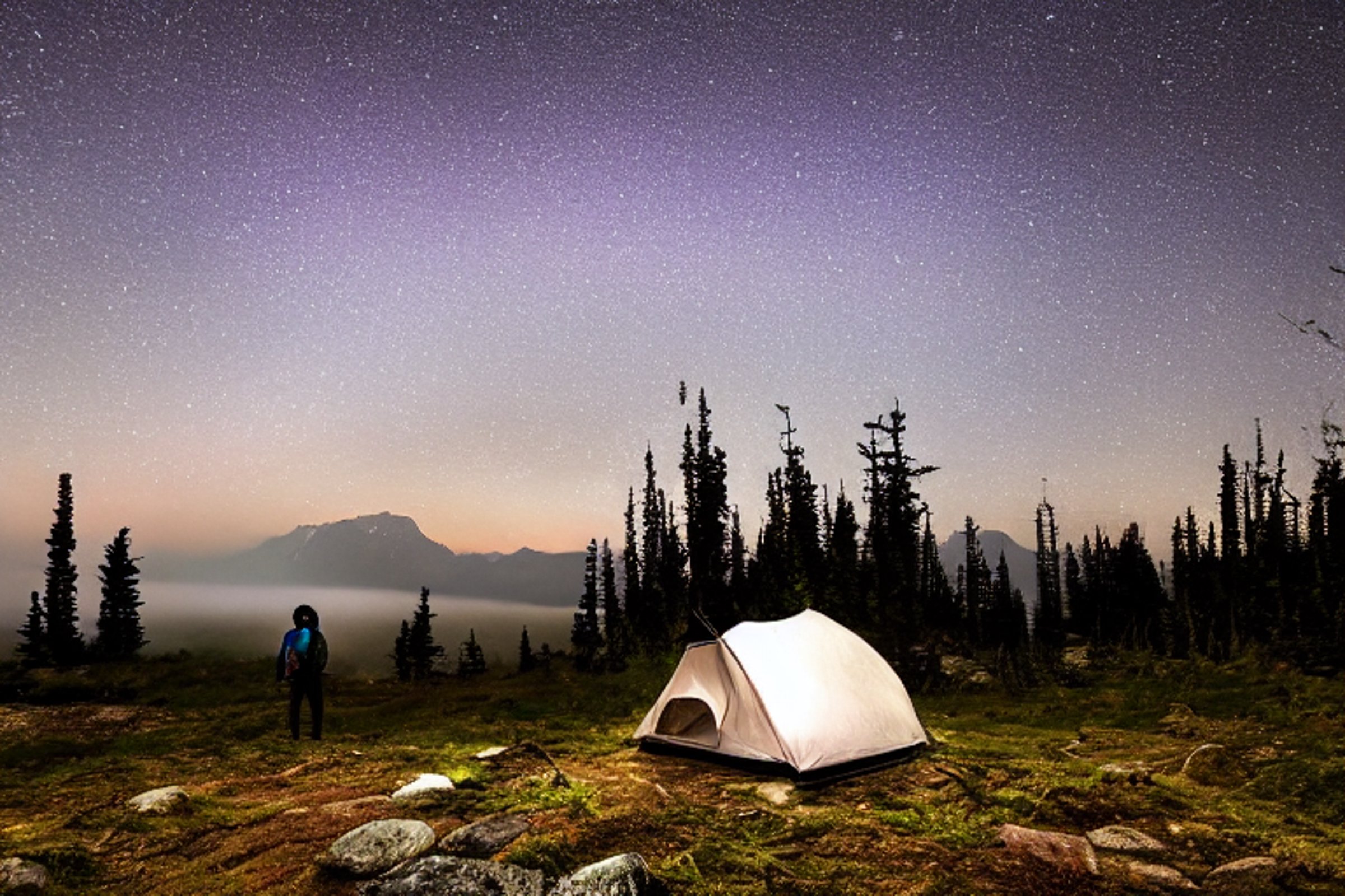 A white tent rests on a rocky dirt campsite beneath glowing green auroras. Small trees and patches of stone surround the foggy camp, creating a quiet, magical night in Washington State.