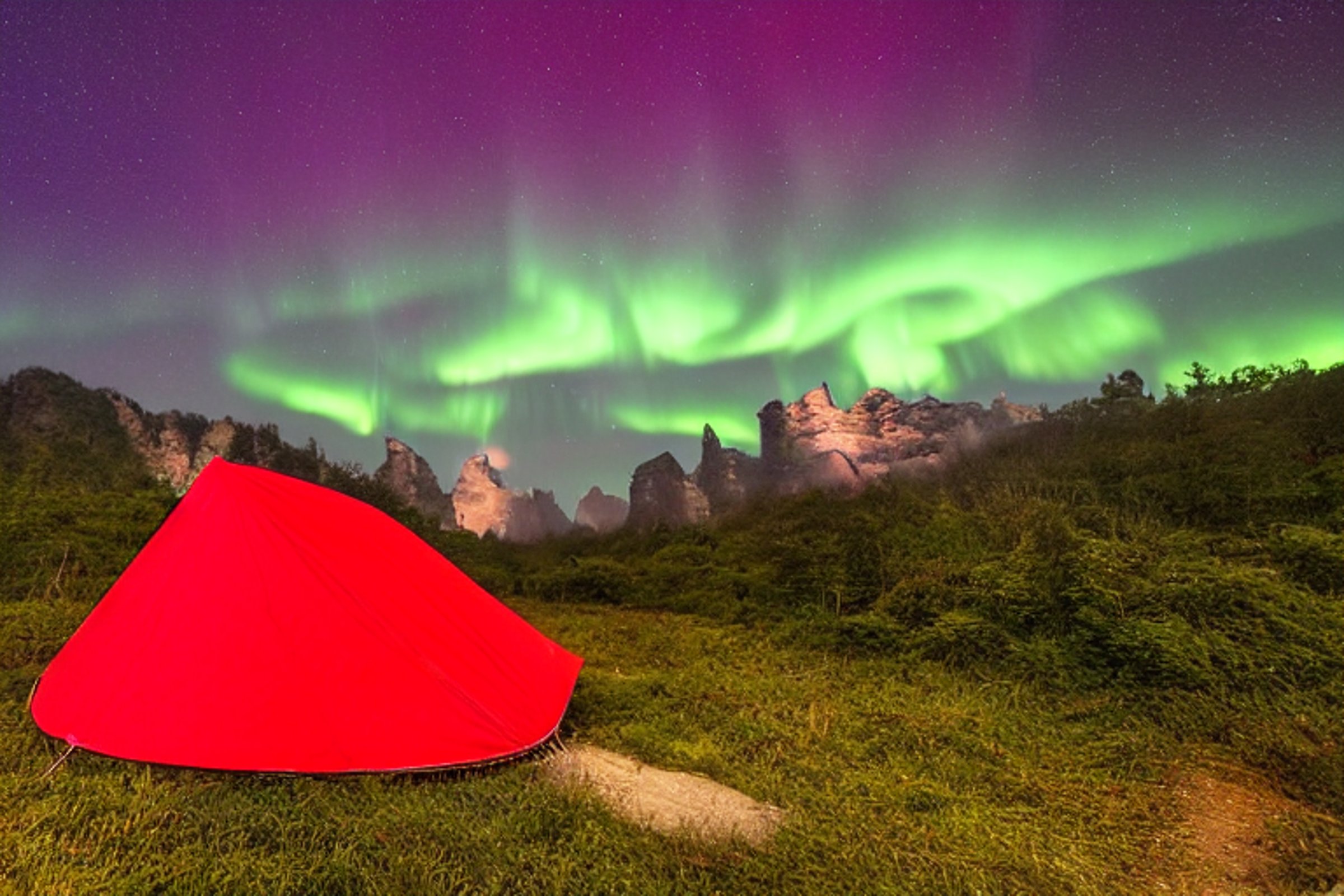 A red and white tent rests on a rocky dirt campsite beneath the glowing northern lights. Tall grass, a winding hiking path, and Zhangjiajie’s wild landscape create a peaceful nighttime setting.