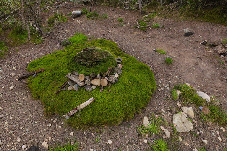 A cozy brown tent rests on a sunny dirt campsite beside a warm hot spring. Mossy trees, stone patches, and a hiking path surround the area, with a large log near the campfire.
