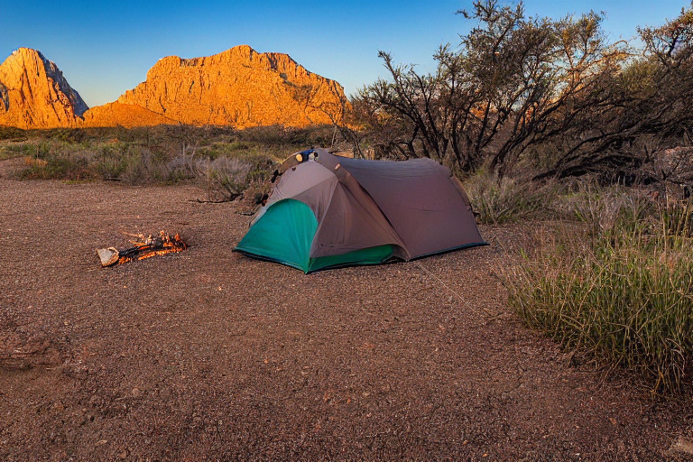 A cozy tent sits beside a glowing campfire in Big Bend National Park. Snow dusts the tall grass and dirt path, while warm golden light fills the cool, quiet campsite. A nearby parking spot makes the peaceful outdoor retreat easy to reach.