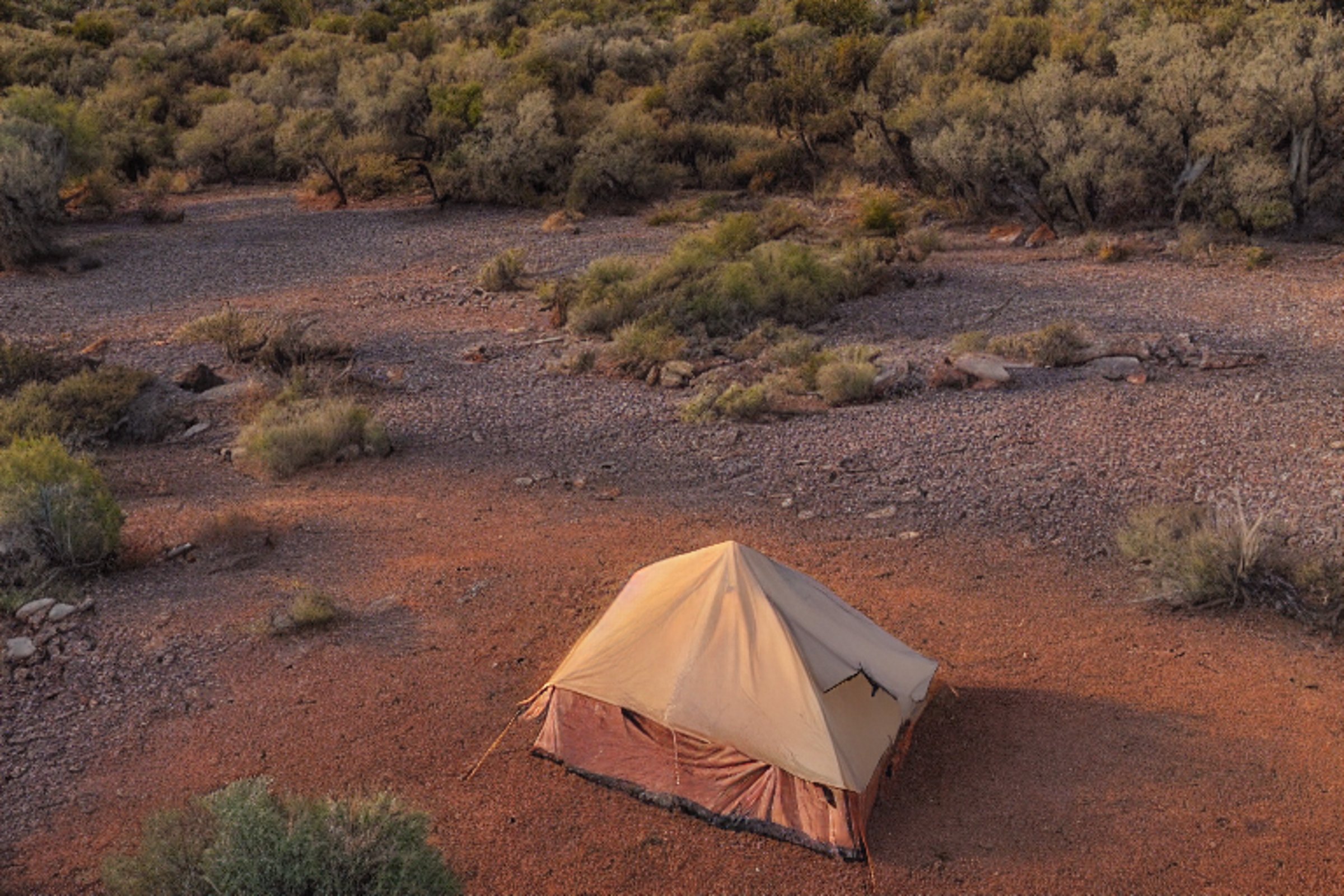 A brown tent rests on a rocky dirt campsite in the New Mexico desert. Large boulders and a wooden log sit near the campfire, while the sunrise fills the clear sky with warm light. A small parking spot completes this peaceful outdoor scene.