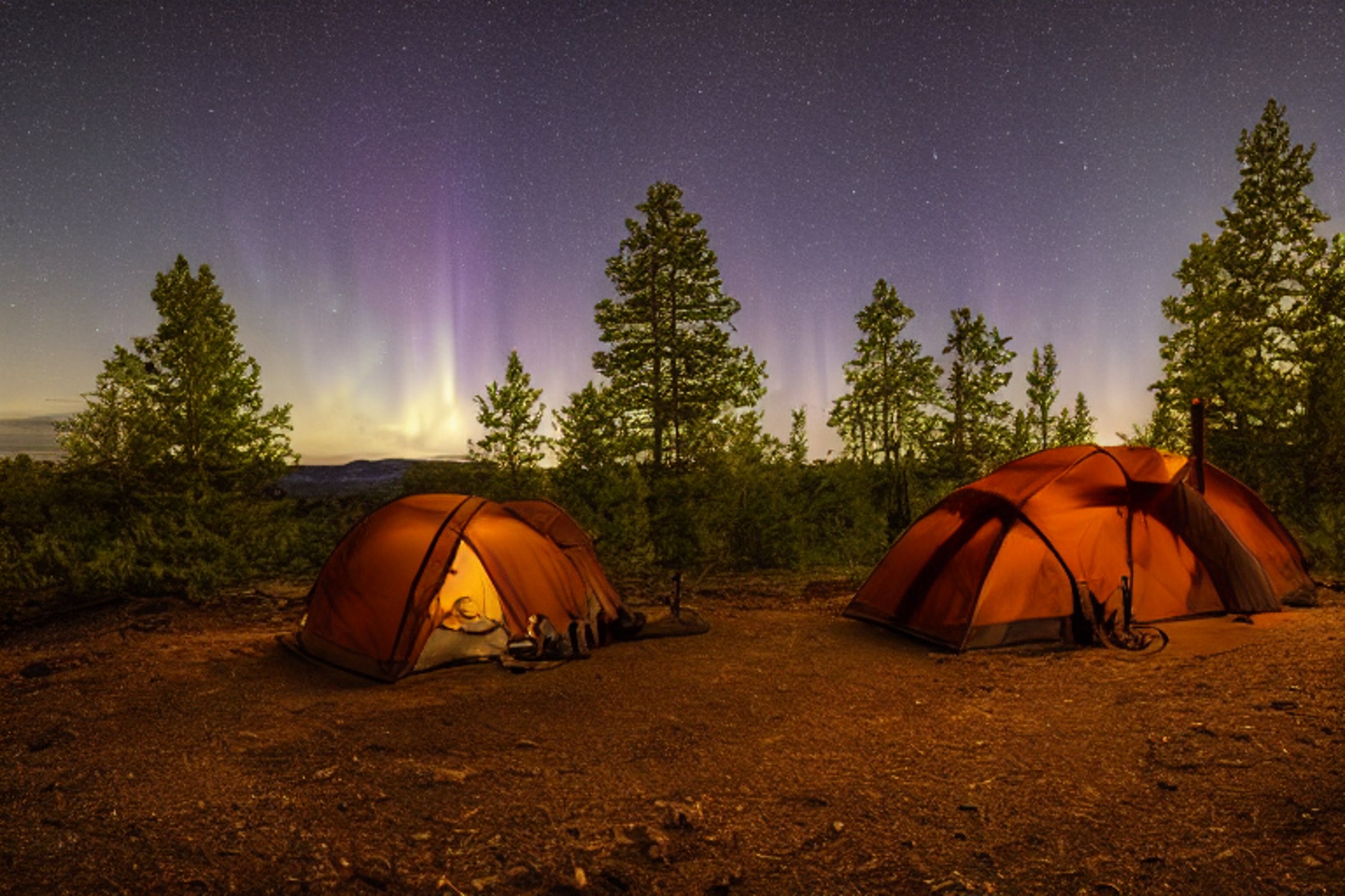 A cozy brown tent rests on a snowy campsite beneath bright green auroras. A hammock hangs between the trees, while a small flashlight glows in the distance.
