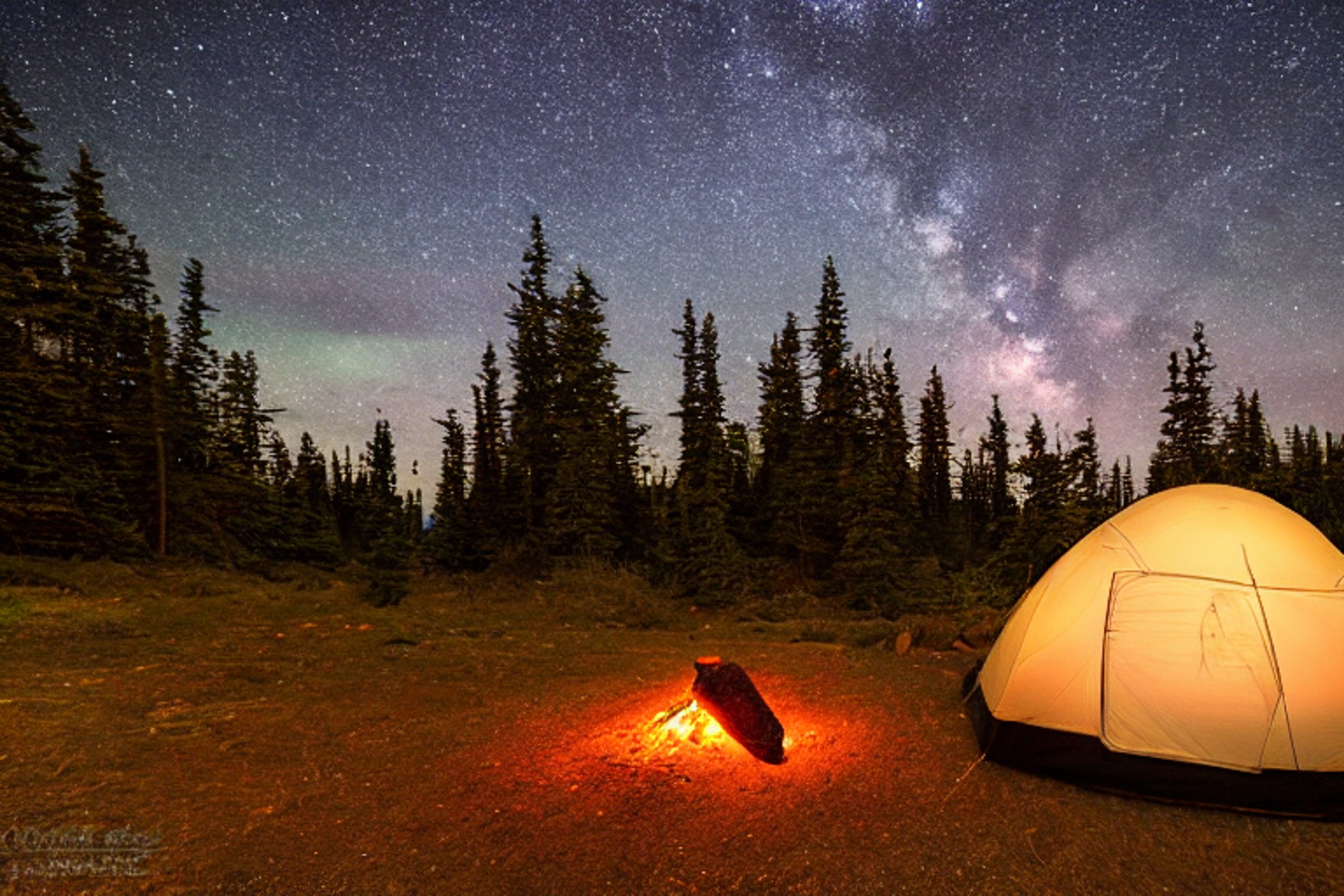 A black tent sits beside a warm campfire on the rocky coast of Maine. Green and purple lights dance above the mountains, with a large rock resting in the rough ground nearby.