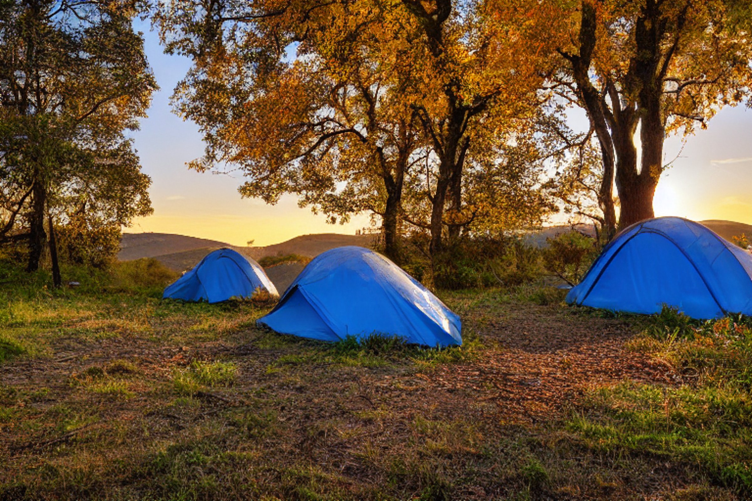 A blue tent rests on a dirt patch at a peaceful riverside campsite. Rolling hills, large boulders, and bright fall trees surround the paved camp area. The warm sunset sky gives the scene a calm, golden glow.